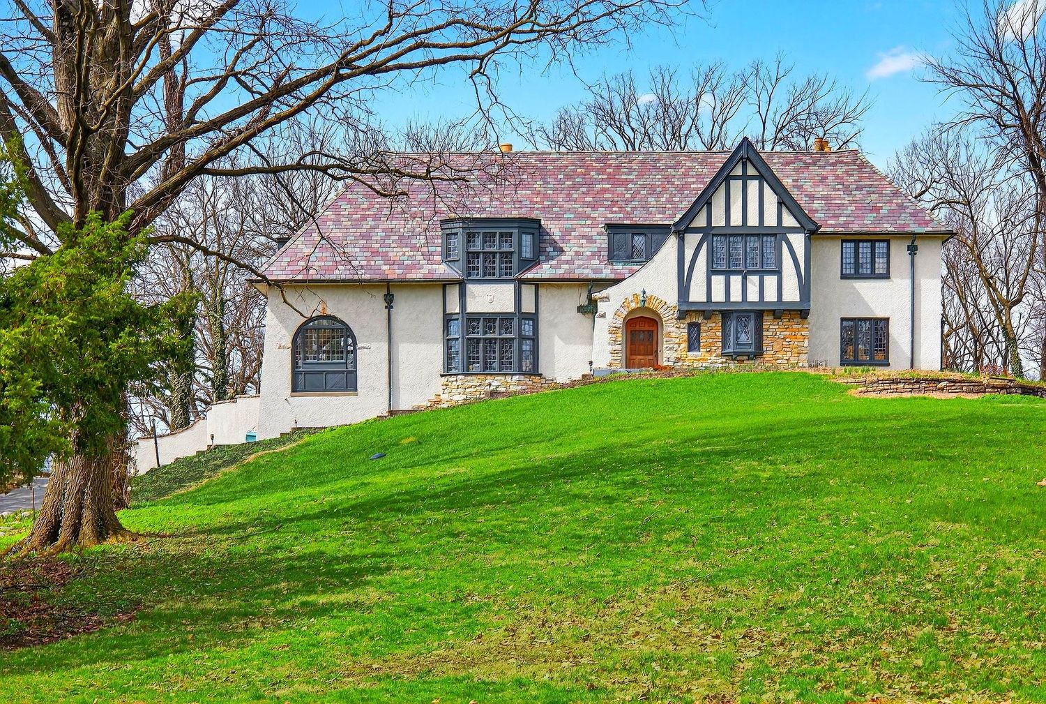 A series of Tudor-style homes with white and brown exteriors sit on rolling green hills beneath bare winter trees.