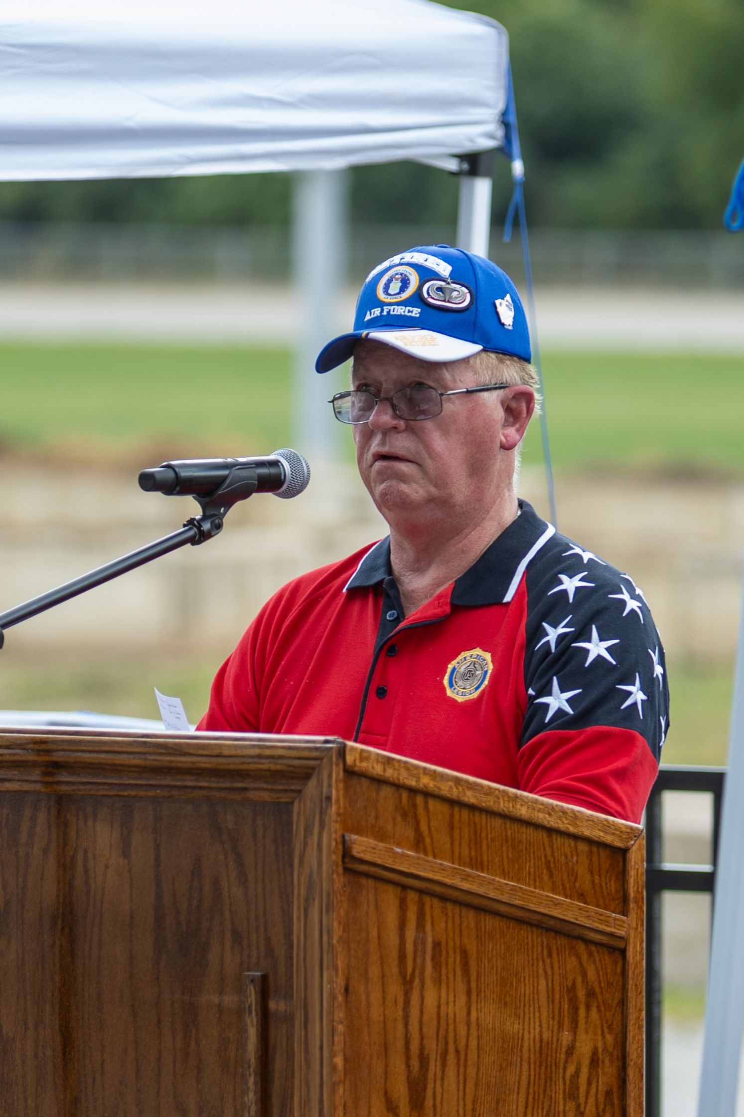 Speaker at wooden podium wearing patriotic American flag-themed attire during outdoor event.