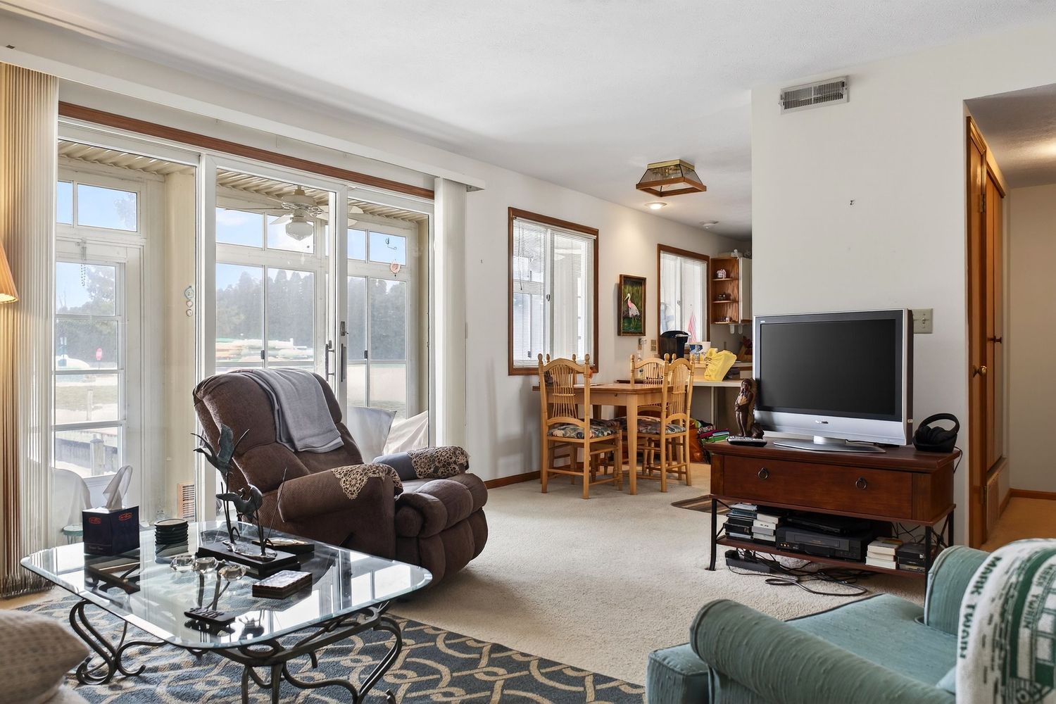 Modern living room with recliner, glass coffee table, TV, and dining area visible through large windows in background.