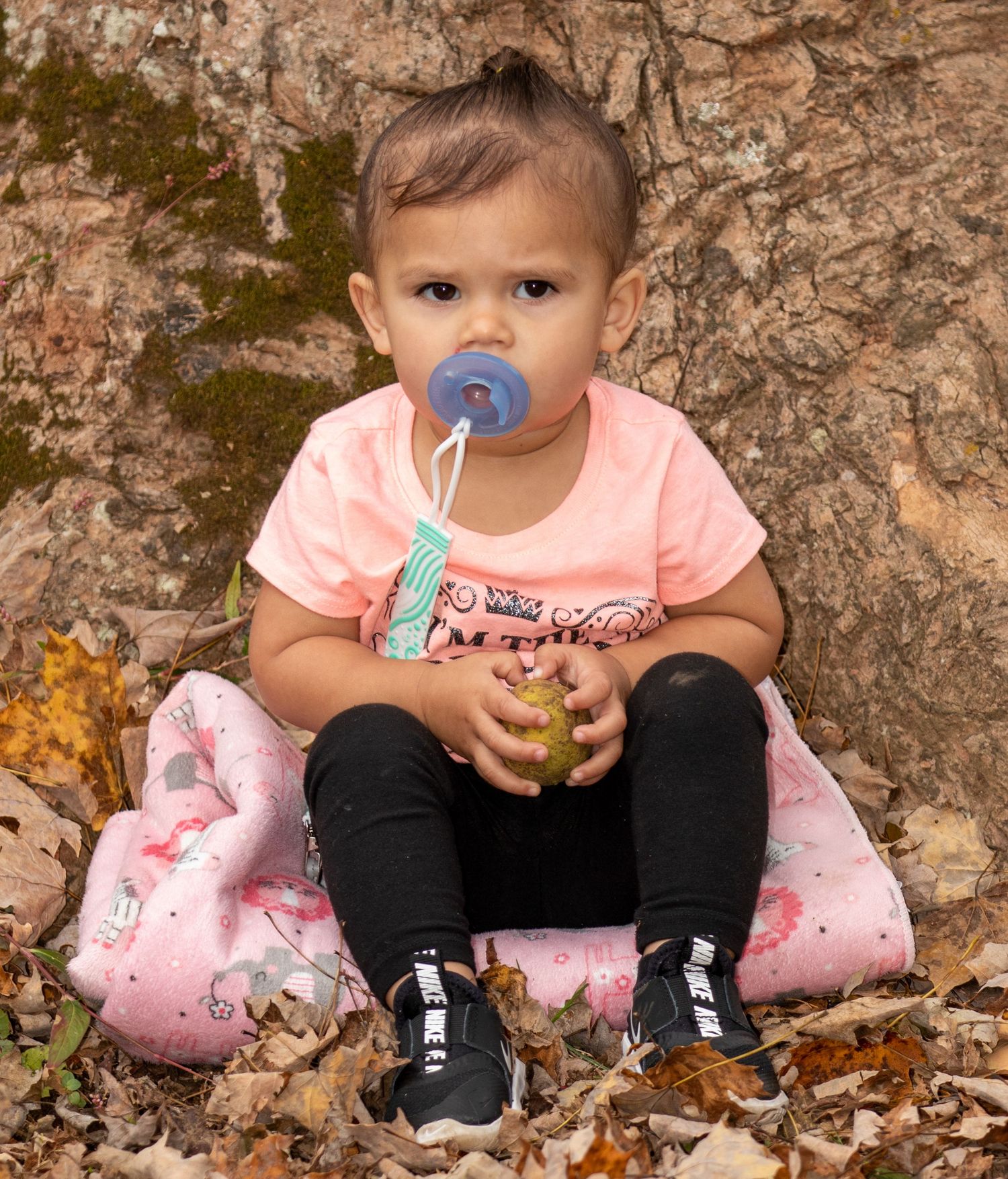 Small child in pink shirt sits on floral cushion with pacifier and holds green item.