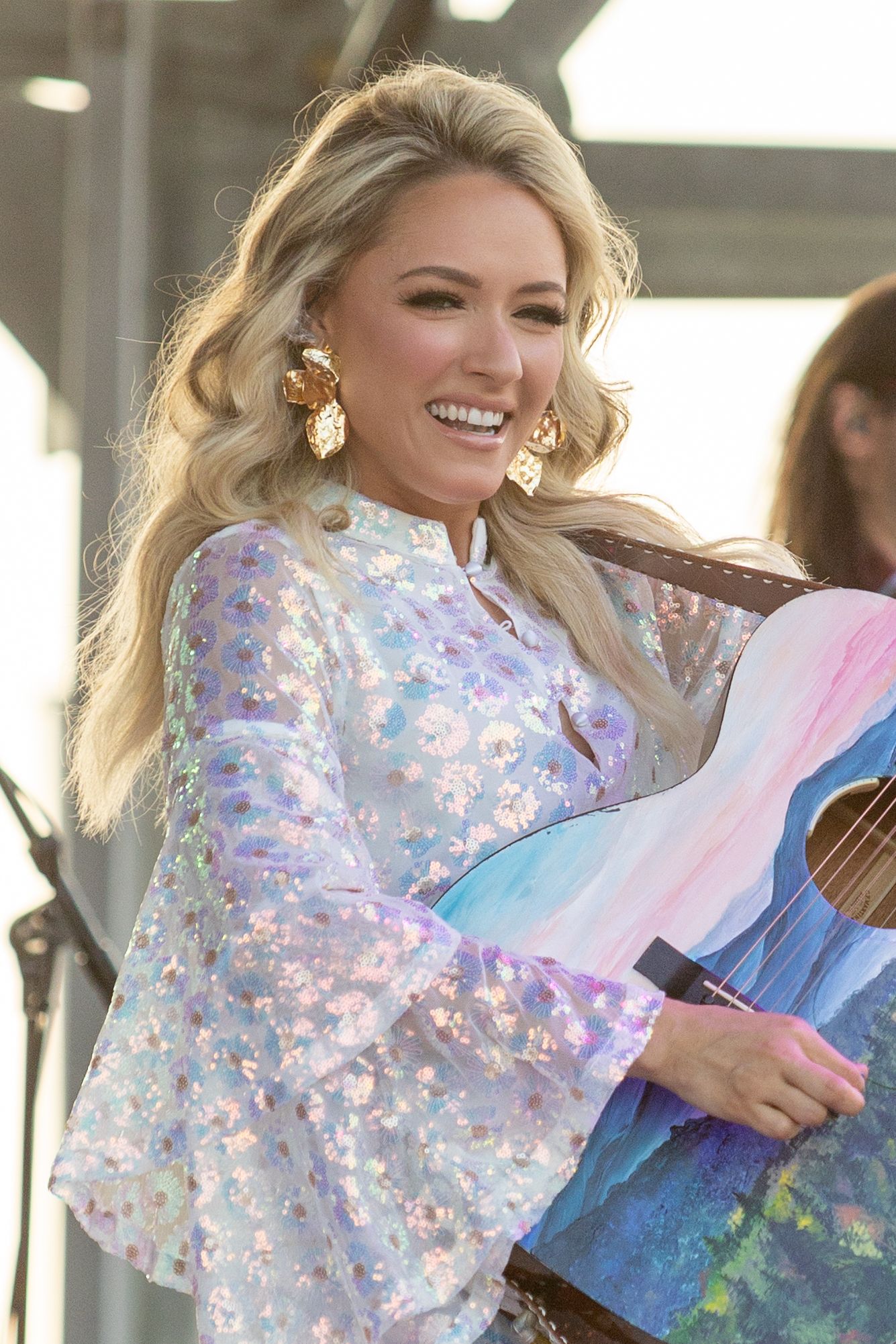 Performer in sparkly white outfit holding colorful artwork on stage.