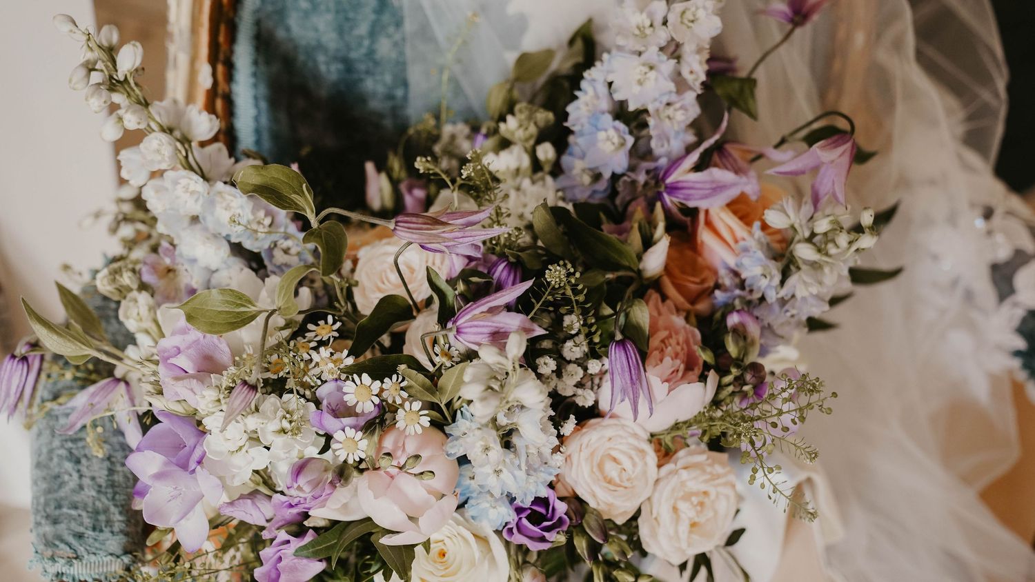 A lush bridal bouquet featuring soft pink roses, white wildflowers, purple blooms, and greenery held near a wedding dress.