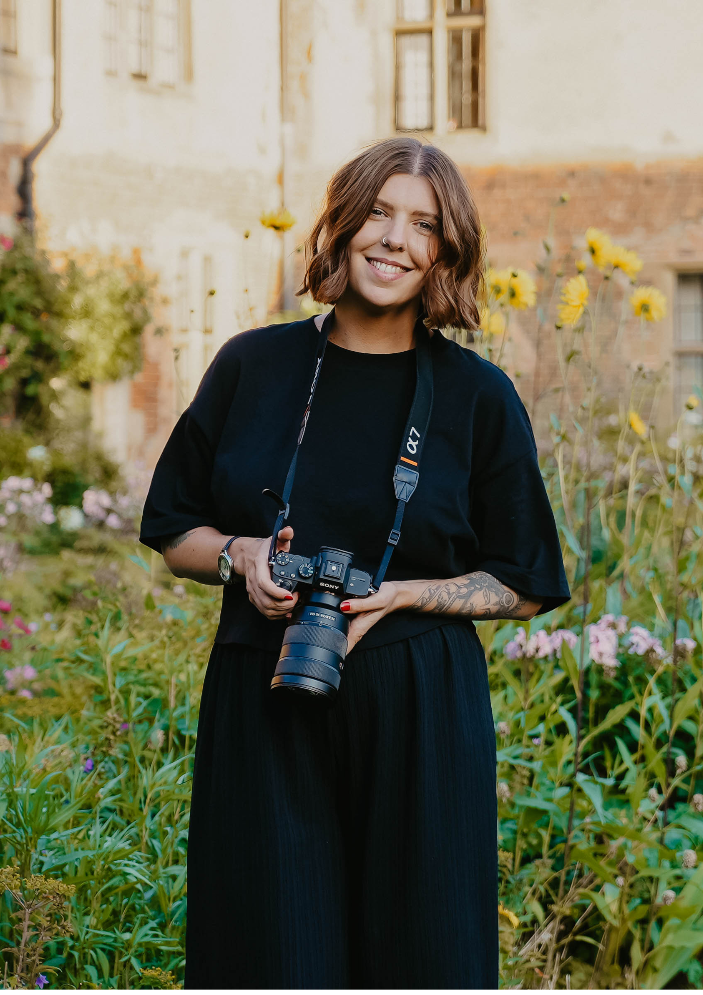 Smiling female photographer in black outfit holding DSLR camera, standing in a lush flower garden.