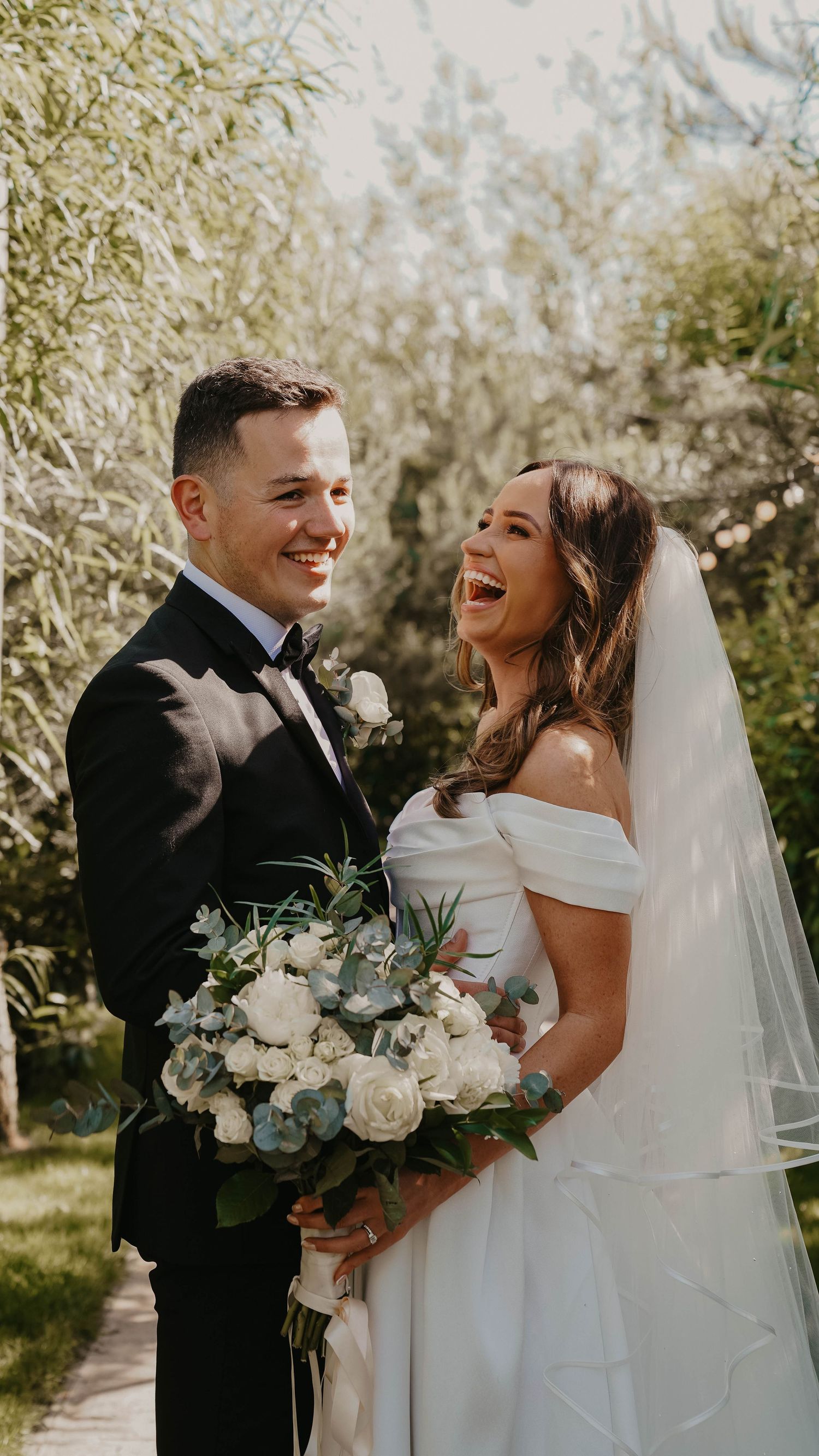 A joyful bride and groom laugh together outdoors, she holds a white floral bouquet and wears an off-shoulder gown with veil.