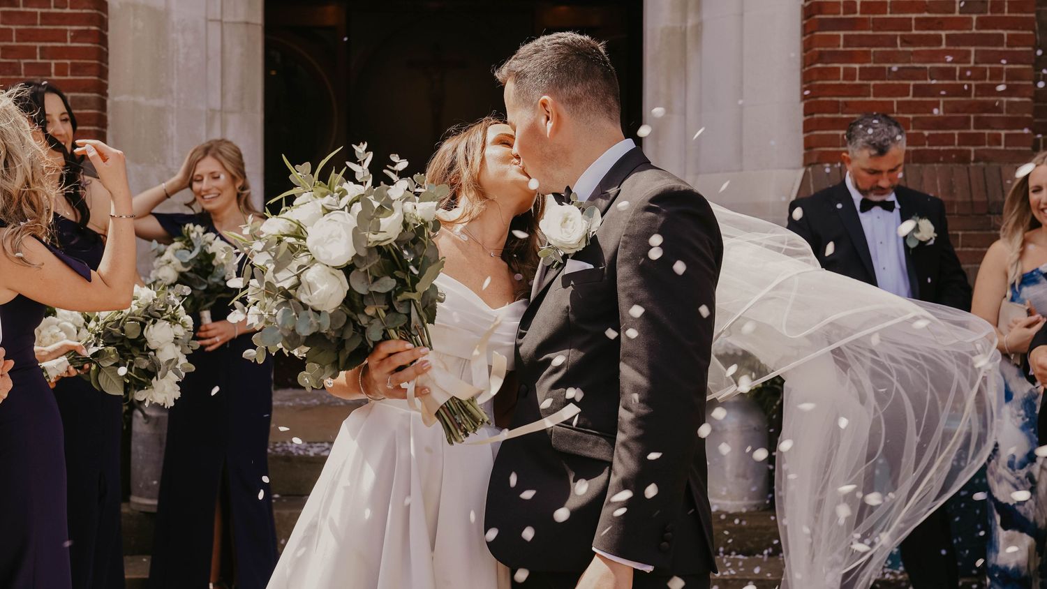Bride and groom share a kiss outside a brick church as guests celebrate with confetti, bride holding a white floral bouquet.