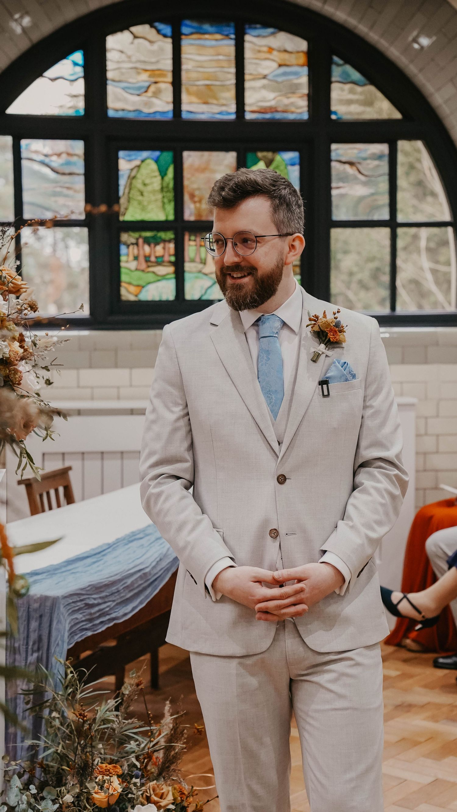 Groom in light beige suit stands in an arched window-lined room with natural lighting.