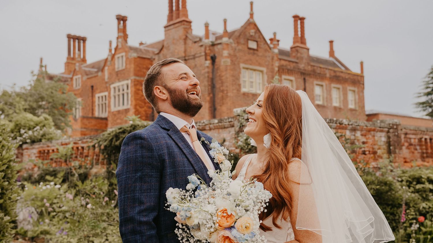 A couple poses in front of a grand Tudor-style brick mansion with ornate chimneys.