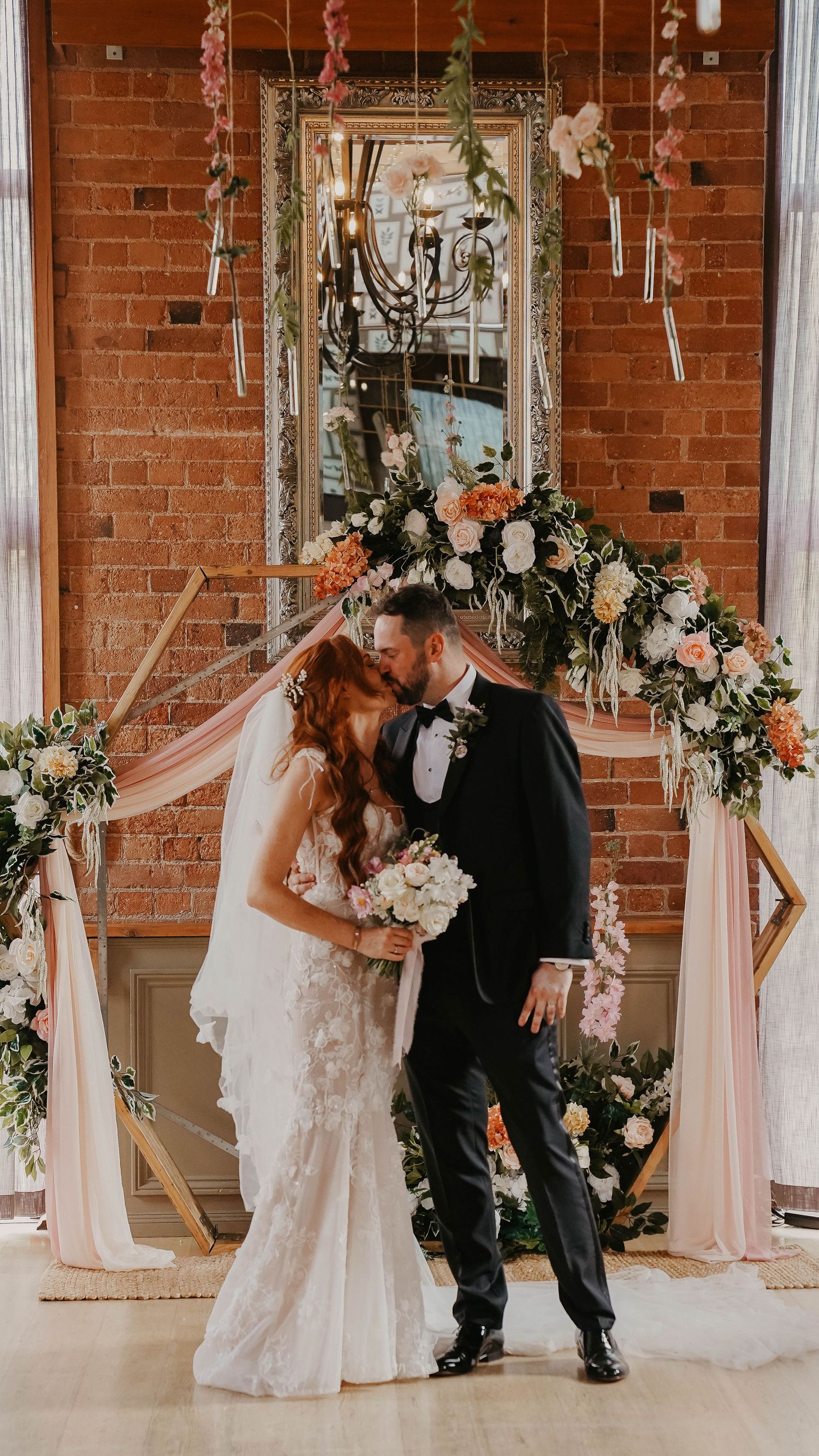 Wedding couple dancing in brick-walled venue decorated with floral arrangements and white draping.