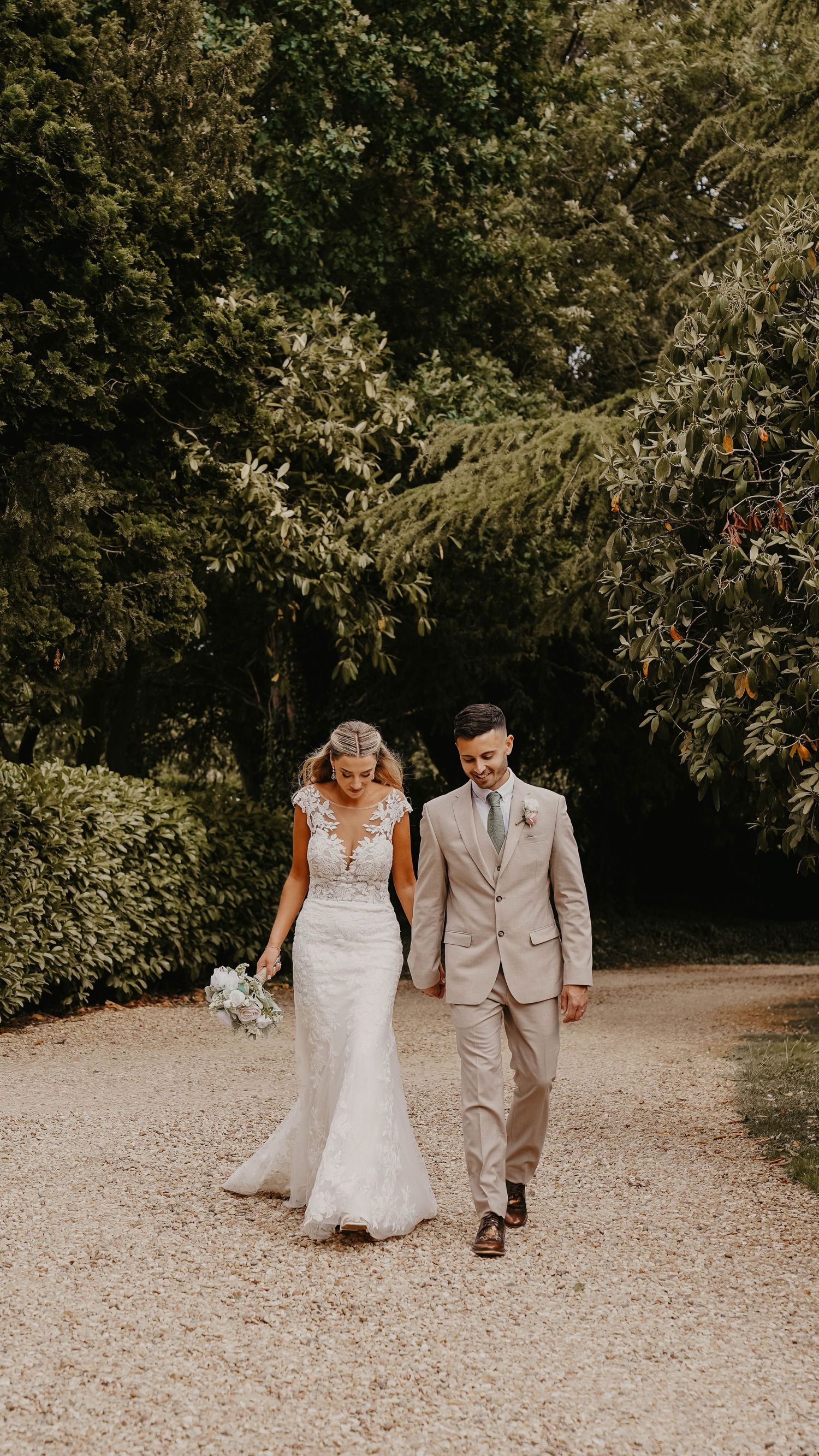 A couple walks hand in hand down a gravel path surrounded by lush greenery.