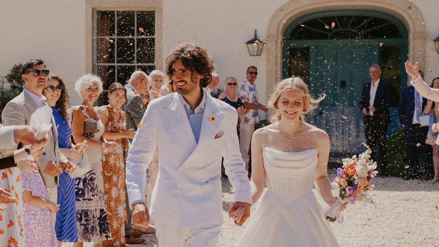 A joyful newlywed couple walks hand-in-hand through a confetti shower, surrounded by cheering guests at an outdoor wedding.