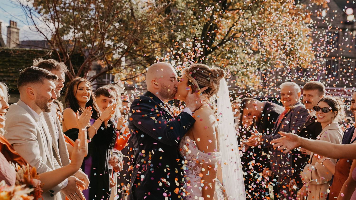 Newlywed couple kissing under colorful confetti shower surrounded by cheering guests at outdoor wedding ceremony.