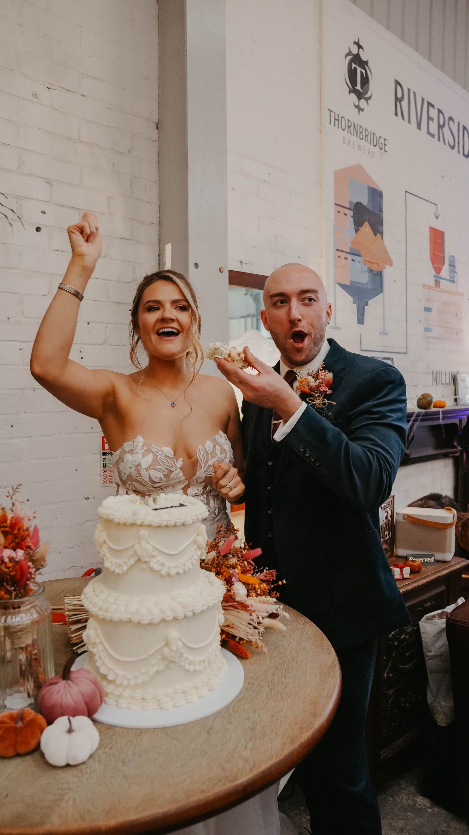 Couple celebrating with enthusiasm next to their white tiered wedding cake.