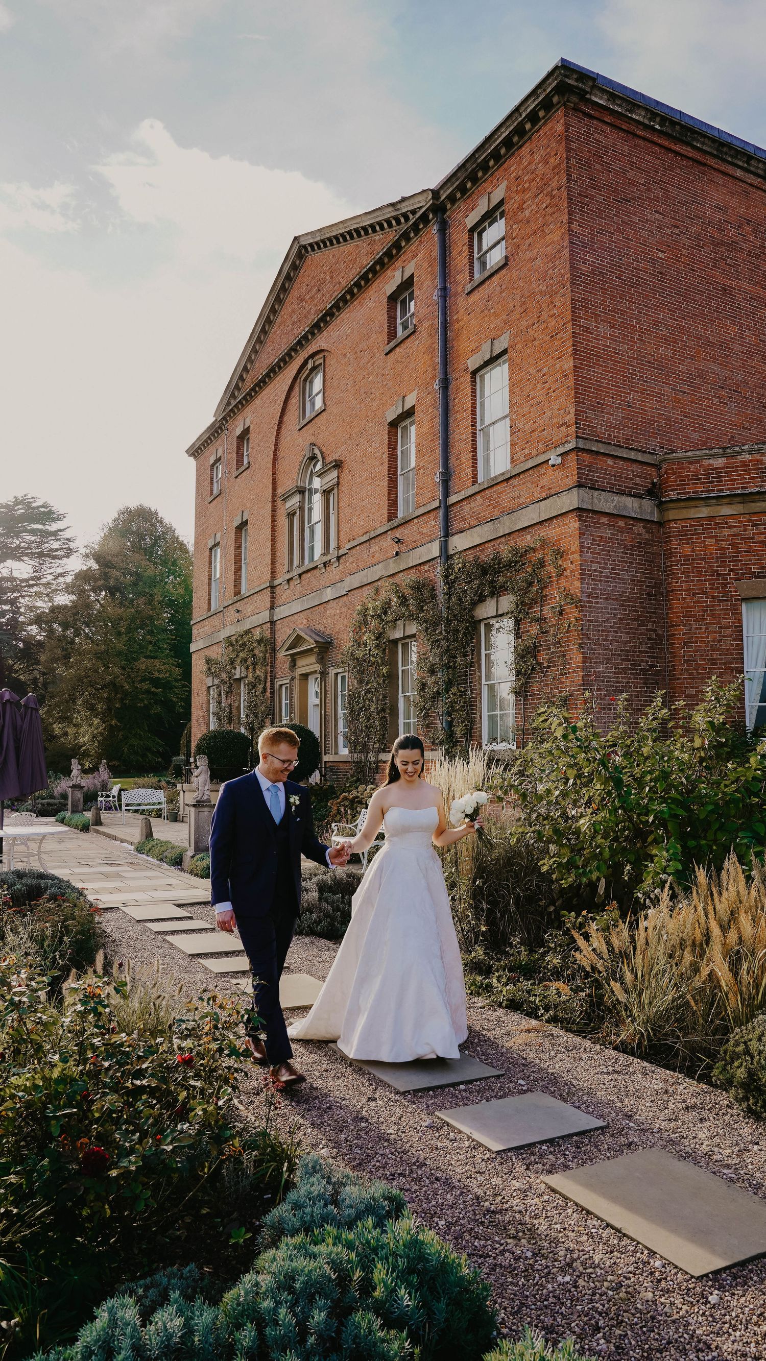 Couple walks along path outside historic brick building during wedding photoshoot.