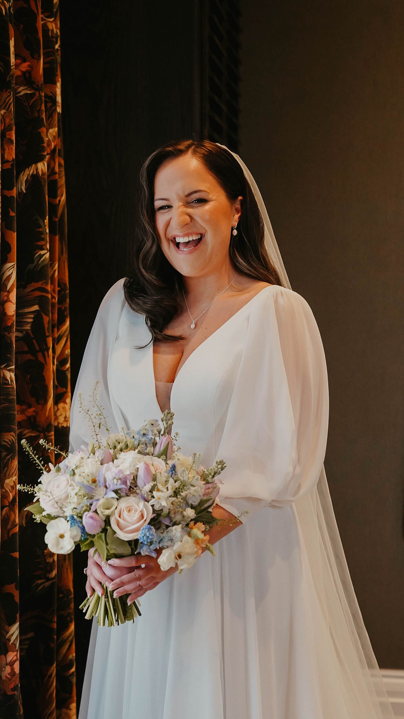 Bride in elegant white dress and veil holds a beautiful pastel bouquet while laughing joyfully.