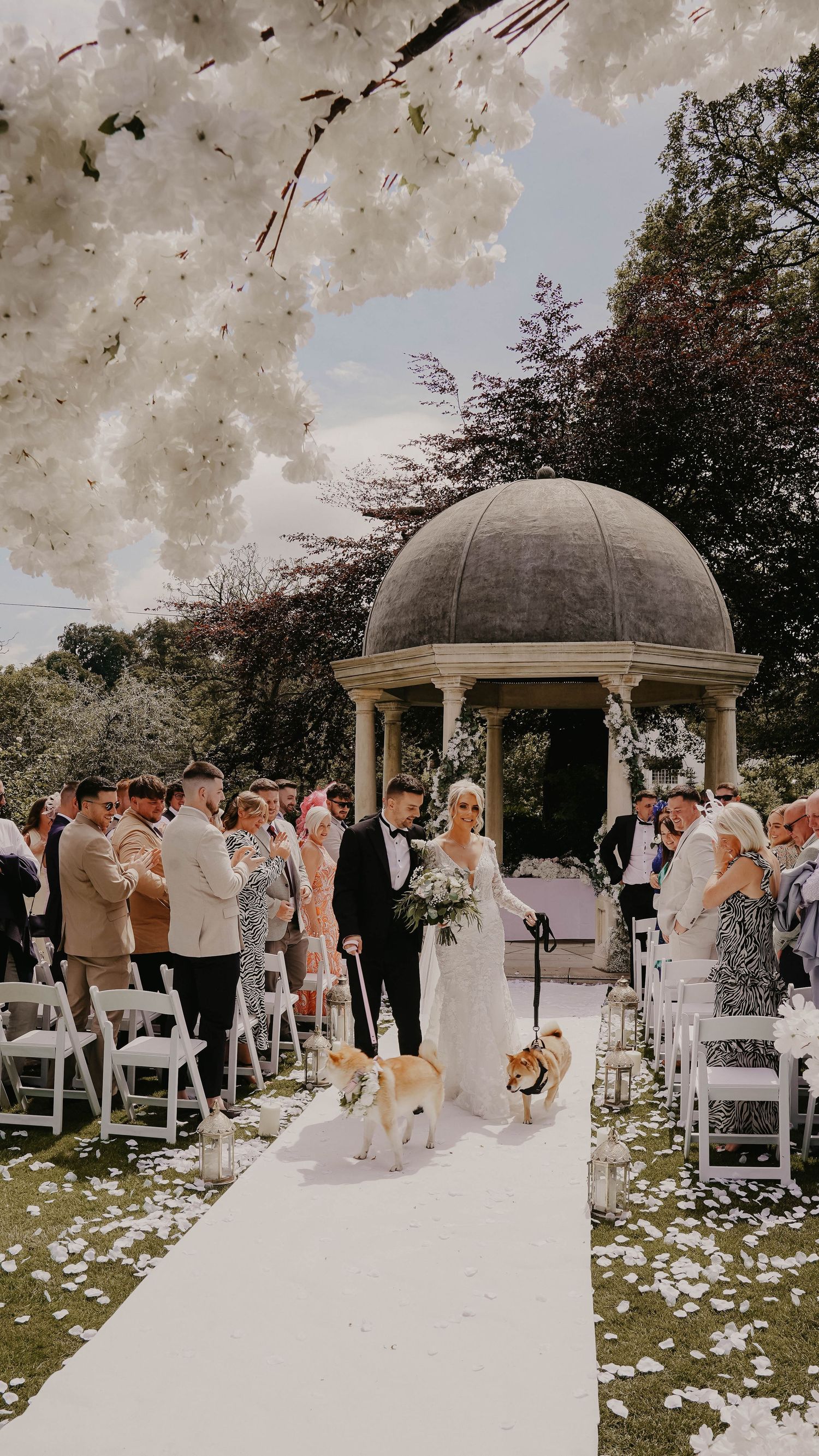 Outdoor wedding ceremony setup with white chairs and domed gazebo surrounded by blooming flowers.