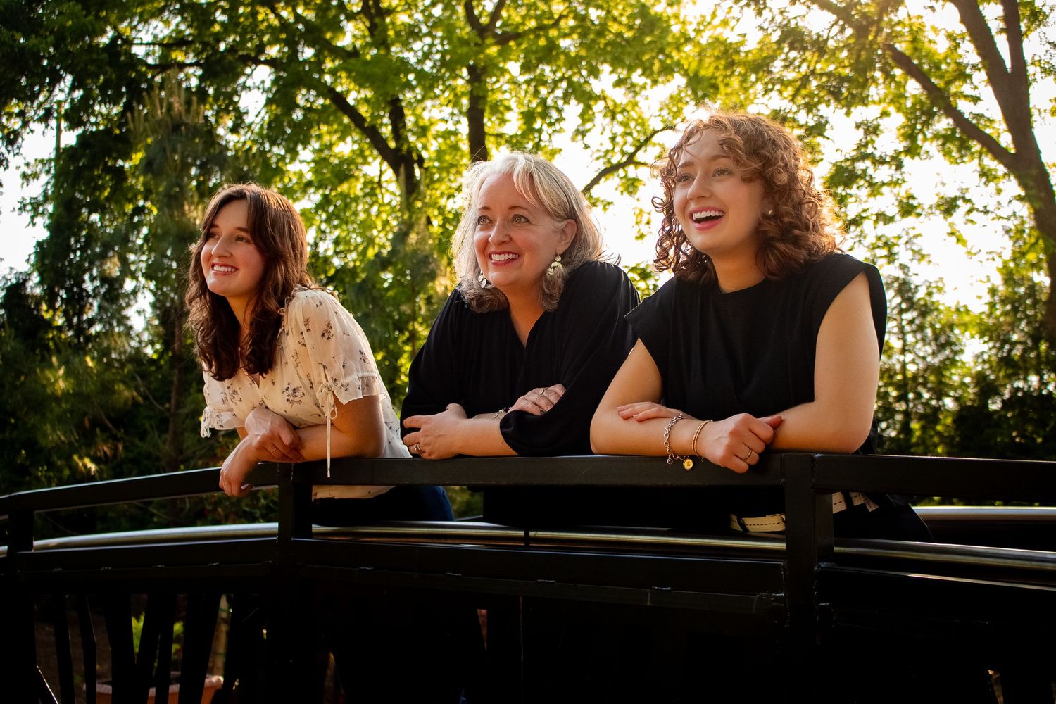 Three women smiling and leaning on a railing outdoors, surrounded by lush green trees in warm sunlight.