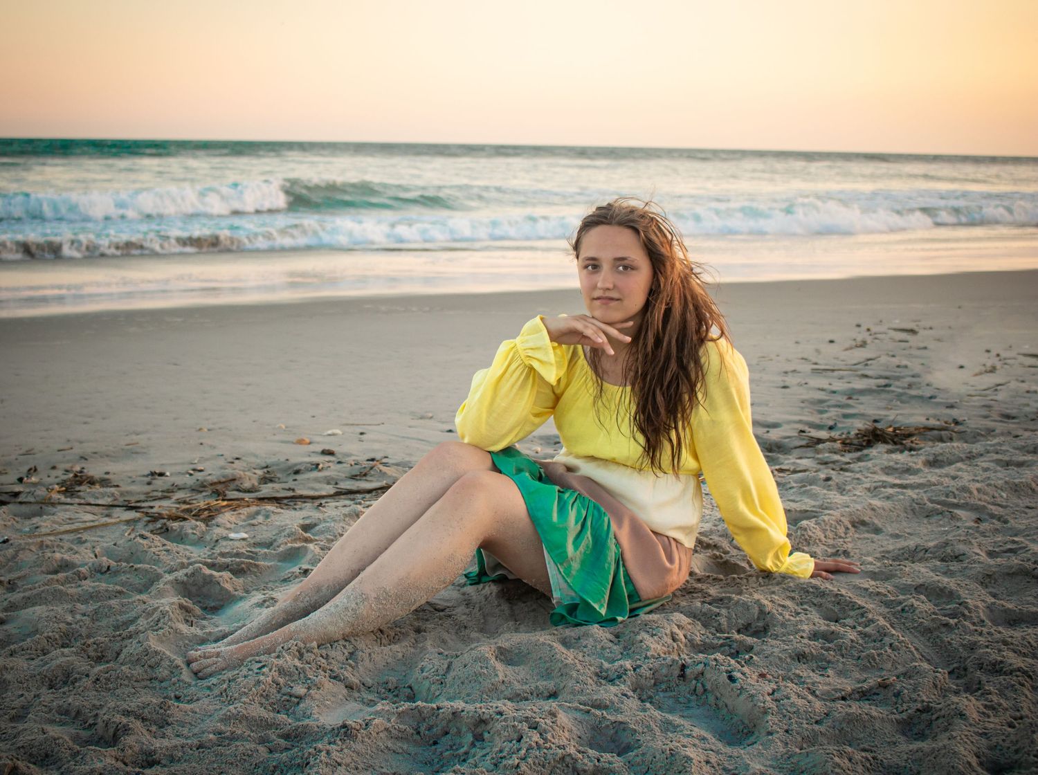 Person in bright yellow top and green shorts sits on sandy beach at sunset with ocean waves in background.