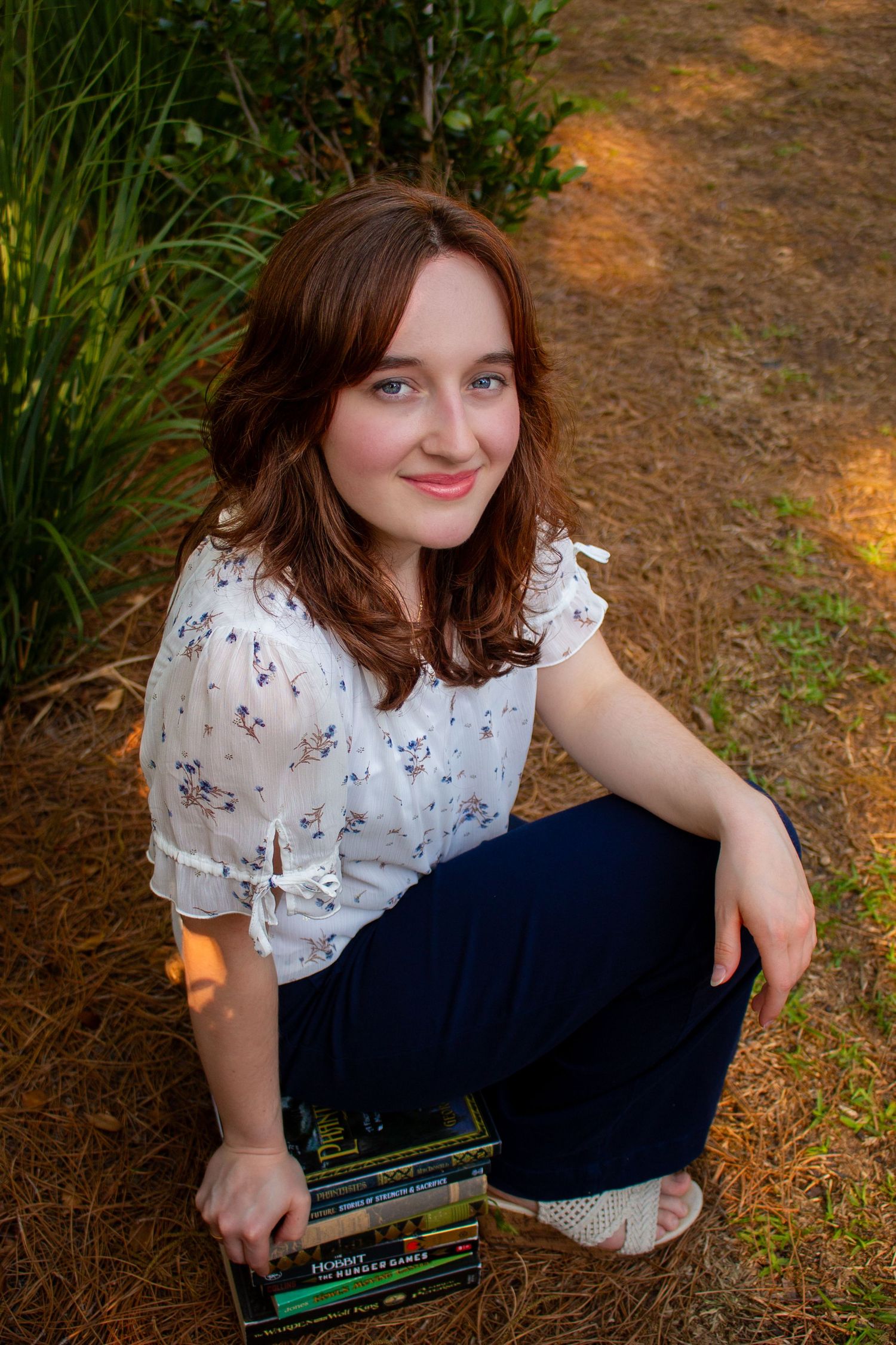 Young woman with auburn hair sits outdoors holding books, wearing floral blouse and navy pants, smiling at camera.