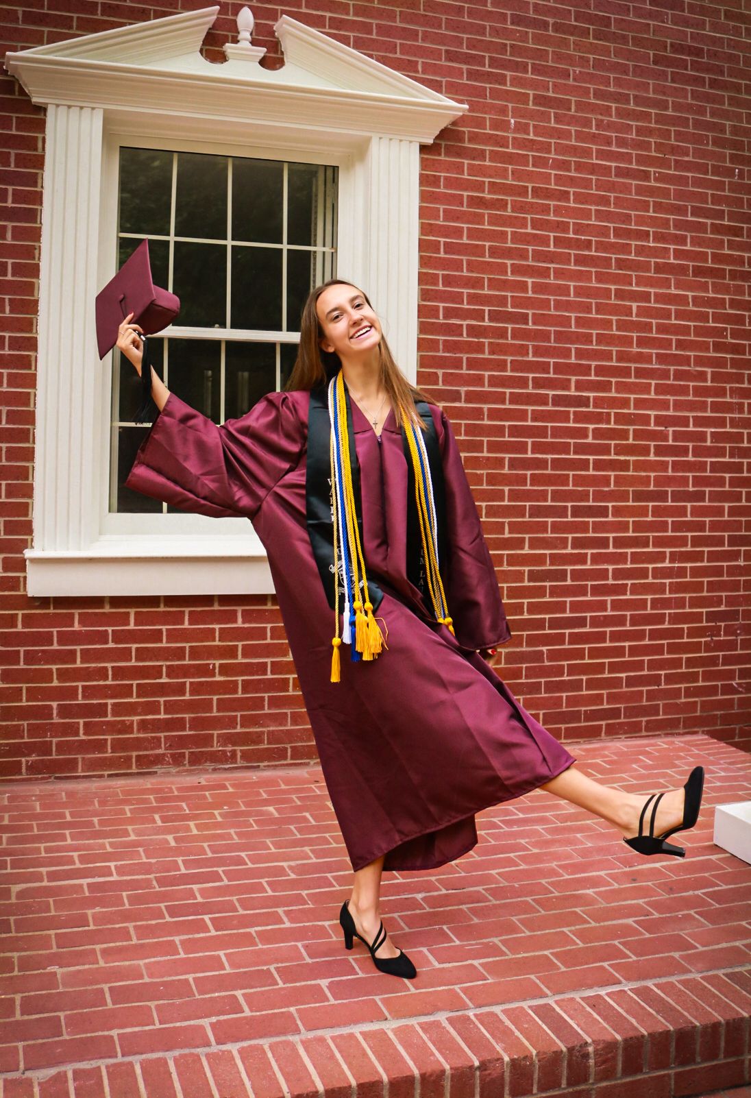 Graduate in maroon cap and gown strikes joyful pose with colorful honor cords against brick building.