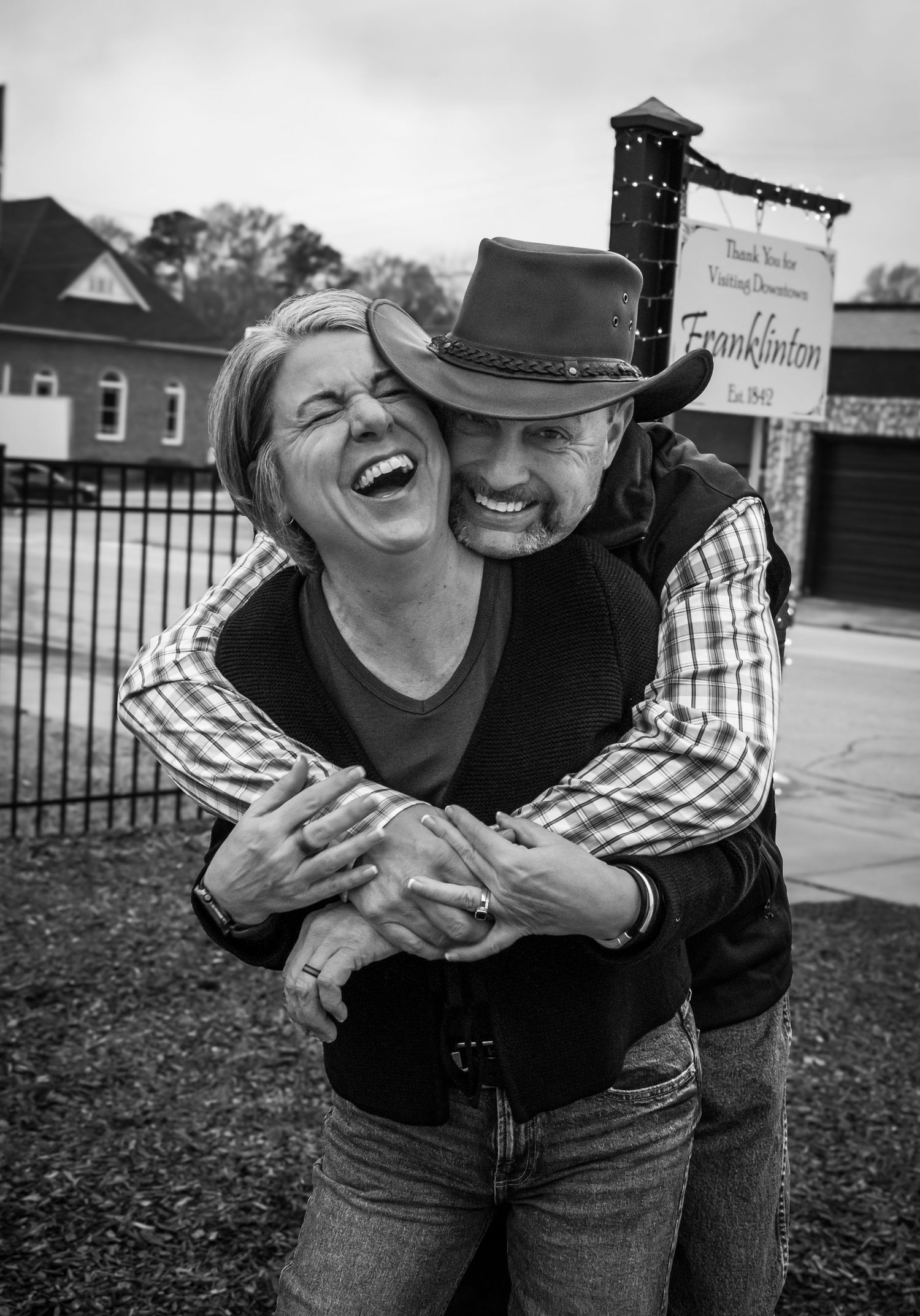 Black and white photo of a laughing couple hugging joyfully in front of a Franklinton welcome sign.