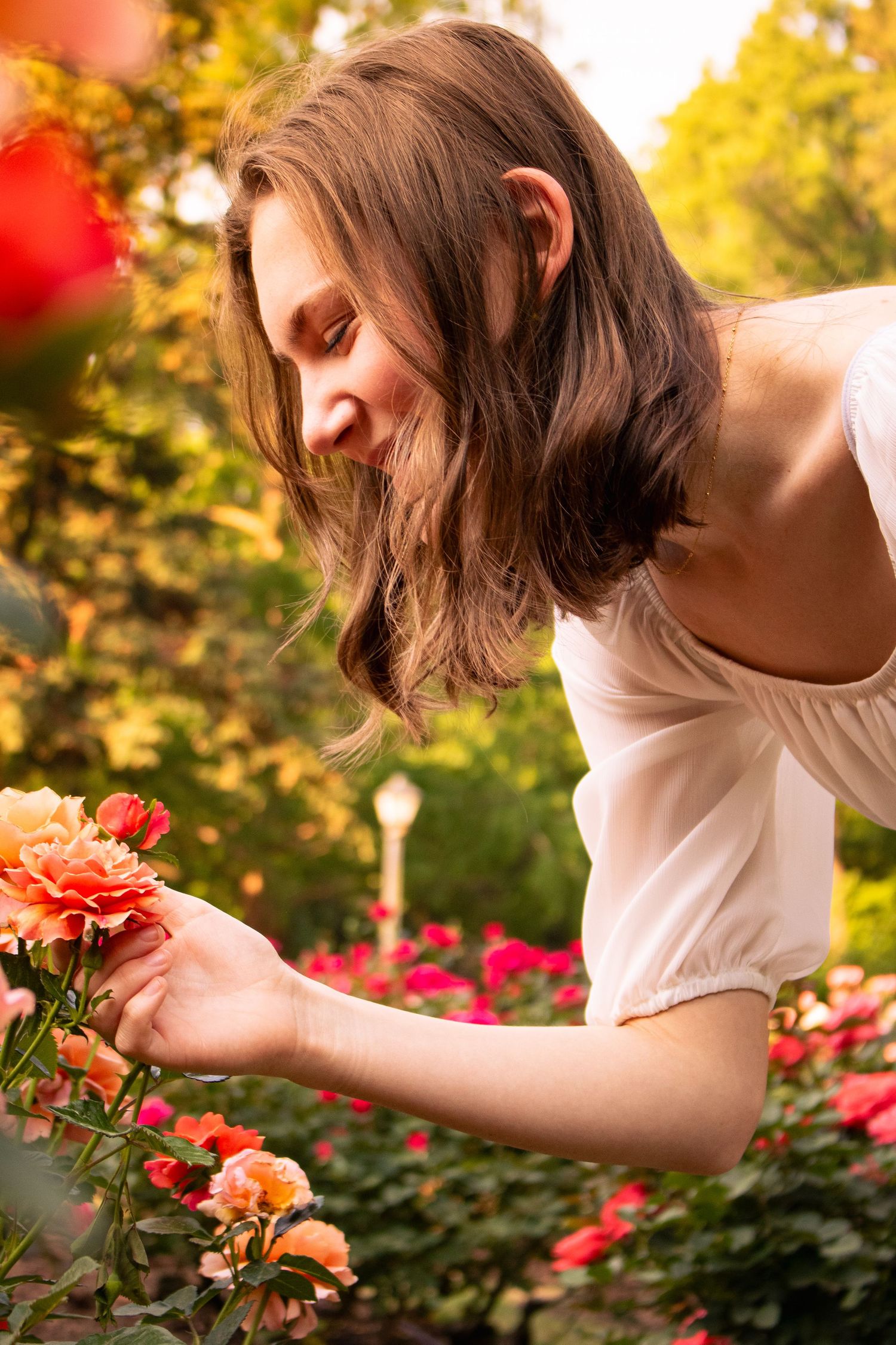 Young woman smiling while smelling orange roses in a lush garden park on a sunny day.