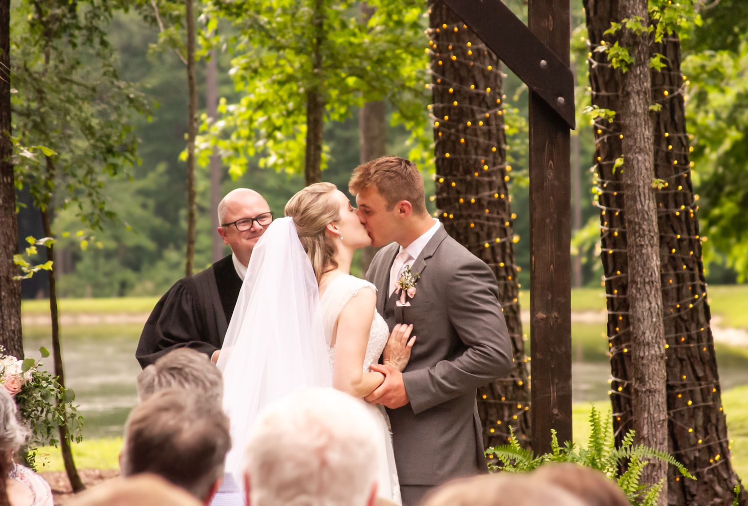 Bride and groom share first kiss at outdoor wedding ceremony near a pond, officiant standing behind them.