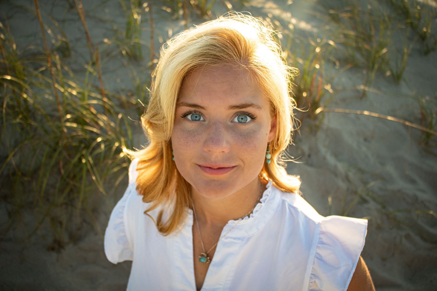 Young blonde woman with blue eyes and freckles in white top, posing at sandy beach dunes in golden hour light.