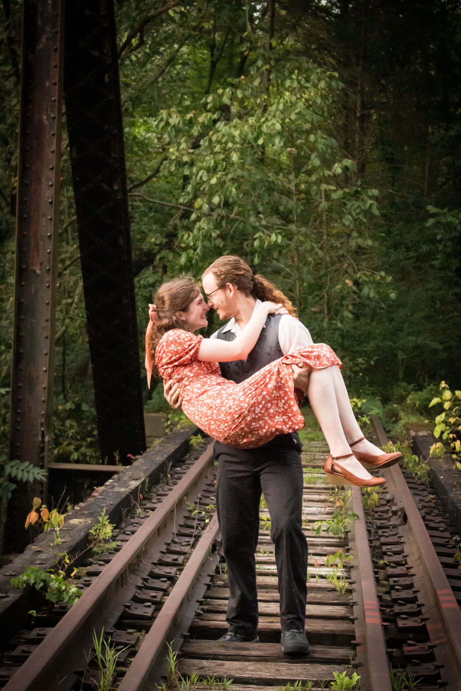 A man carries a woman in a red floral dress on a rustic railway track surrounded by lush green forest trees.