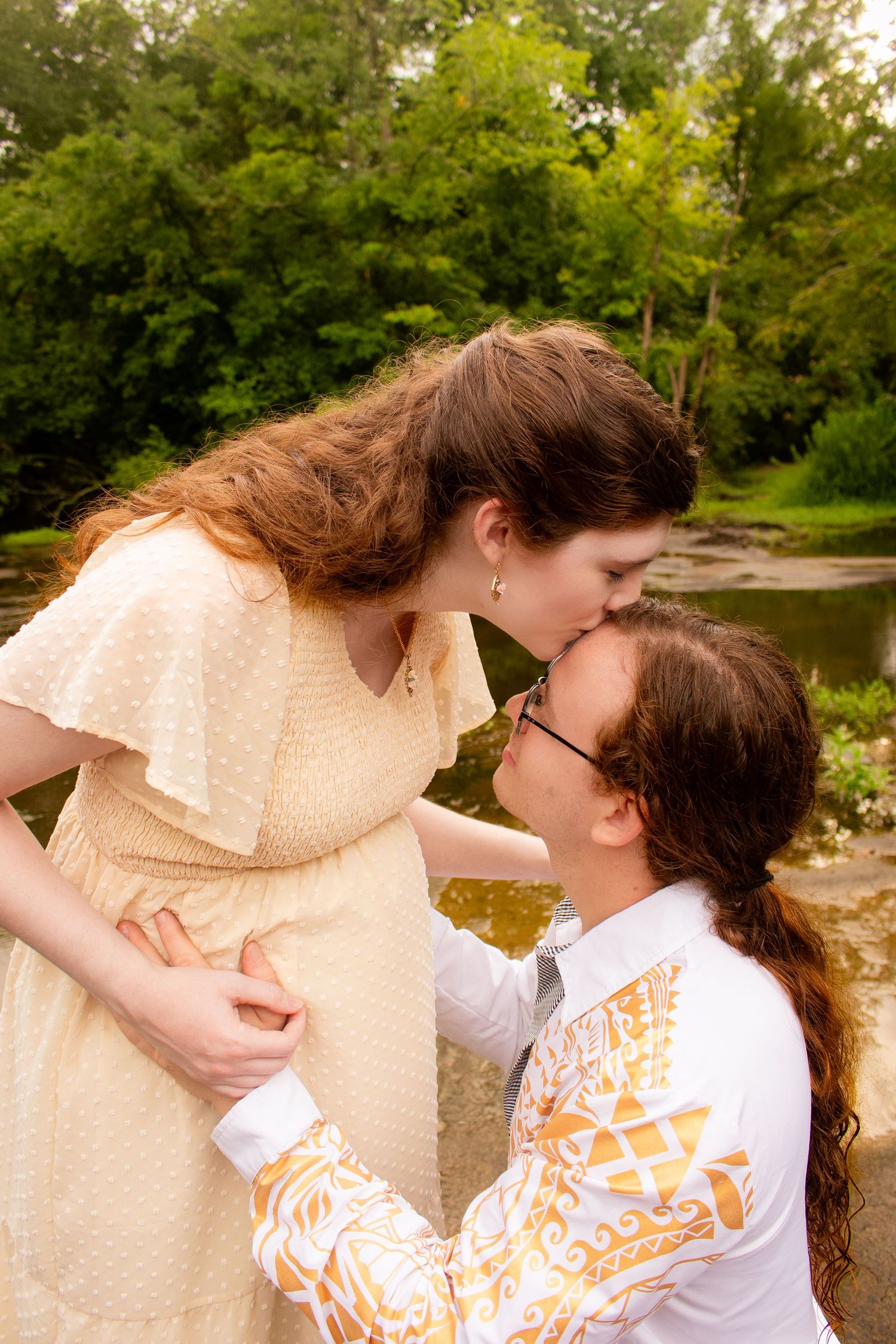 Couple sharing a tender kiss outdoors near a serene river, surrounded by lush green trees in a romantic setting.