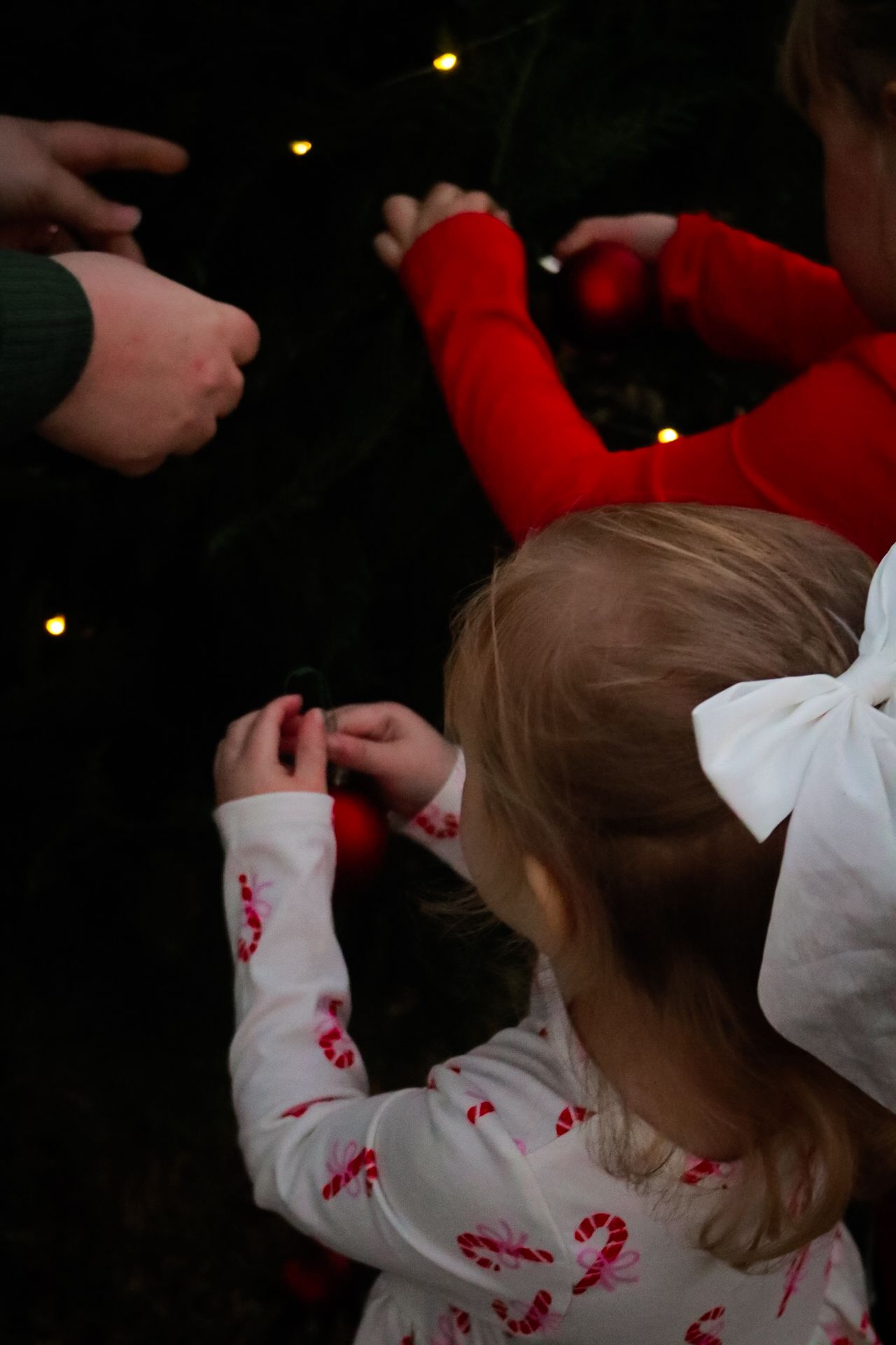 Children in holiday pajamas decorating a Christmas tree with ornaments, with twinkling lights in the background.