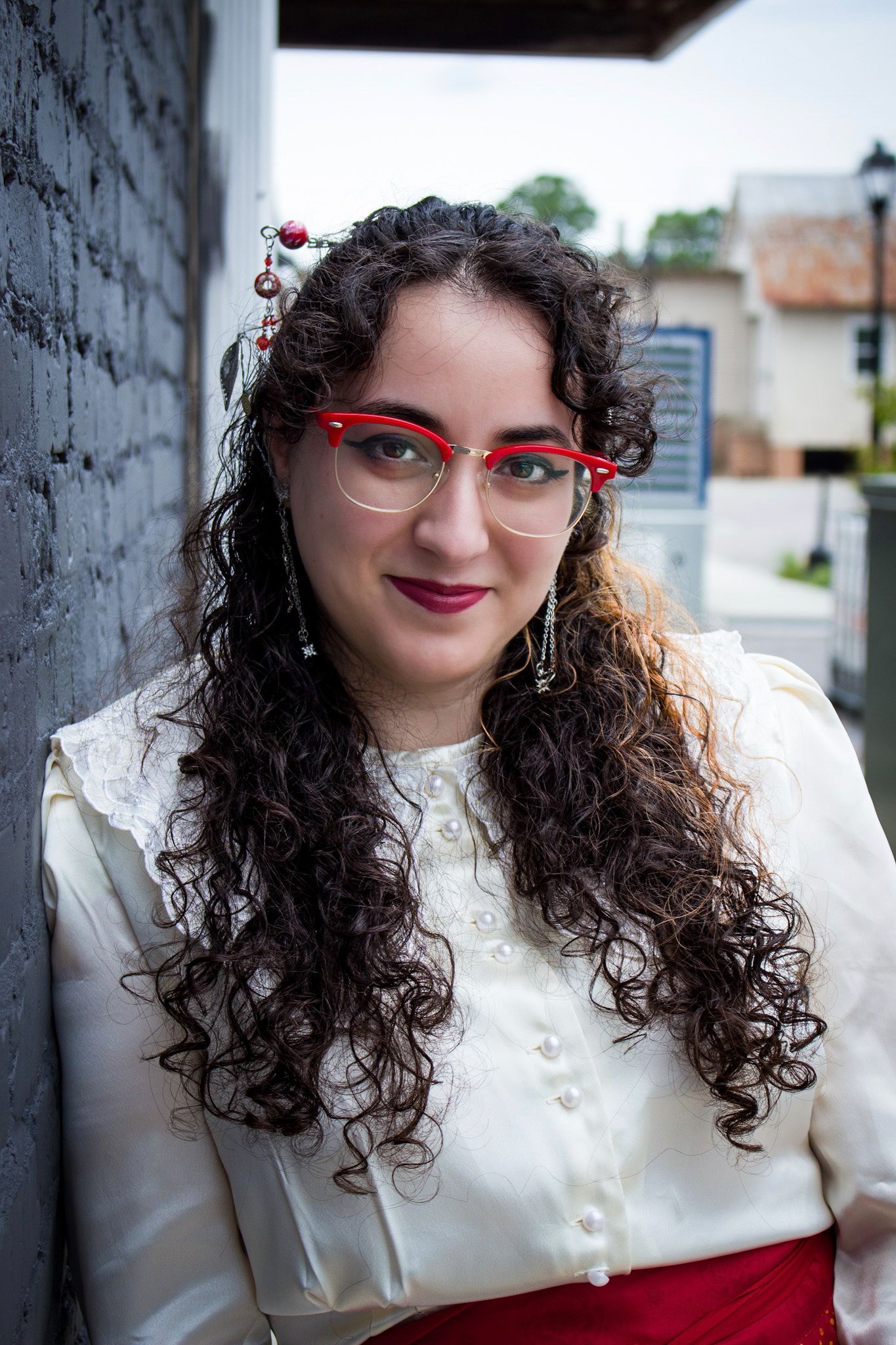 Young woman with curly hair, red cat-eye glasses, and white blouse smiles outdoors near a brick wall.