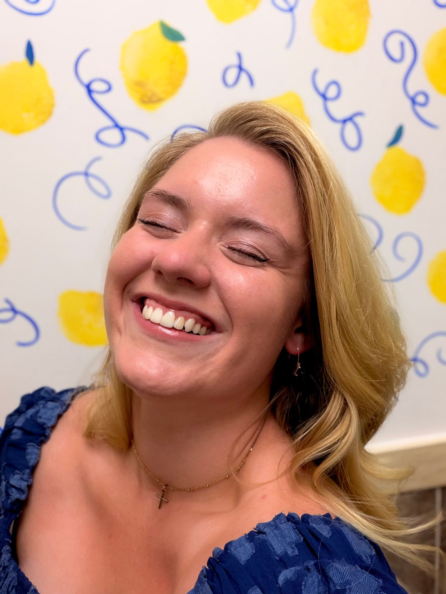 Smiling blonde woman in blue top laughing joyfully against a cheerful lemon-printed wallpaper background.
