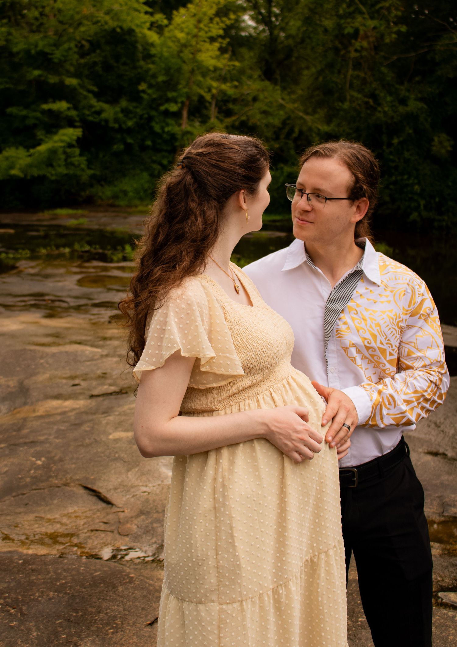 Pregnant woman in cream dress with partner outdoors on rocky riverbank surrounded by lush green trees.
