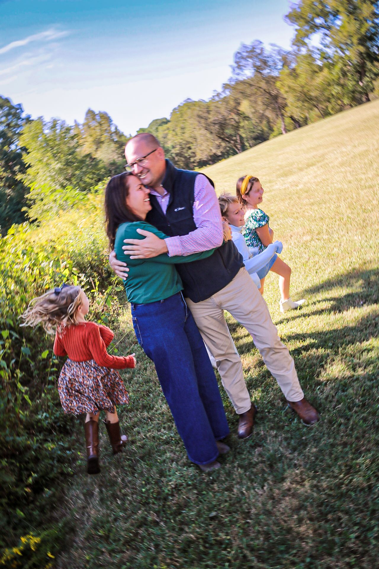 Happy family embracing outdoors in a sunny green field, parents hugging while two young girls play around them.