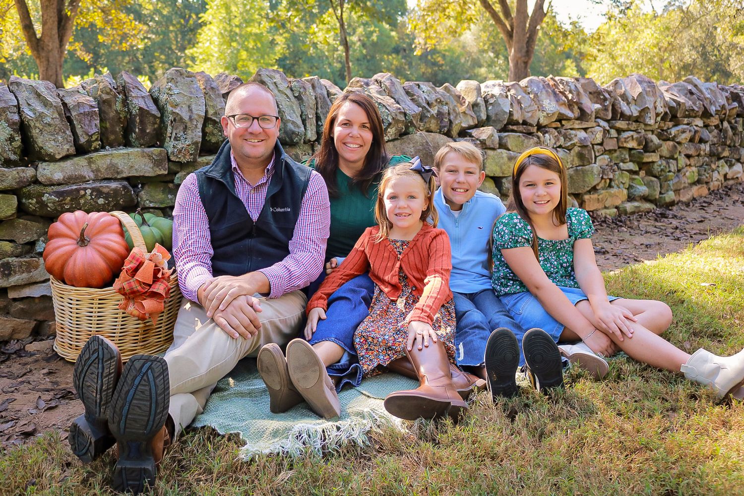 Family of five smiling outdoors in fall setting with pumpkins and stone wall backdrop.