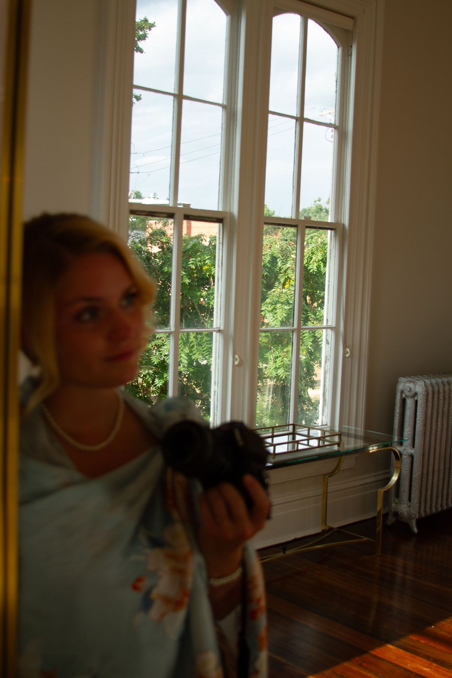 A young girl wearing a light-colored dress stands in front of a large window with a view of greenery outside.