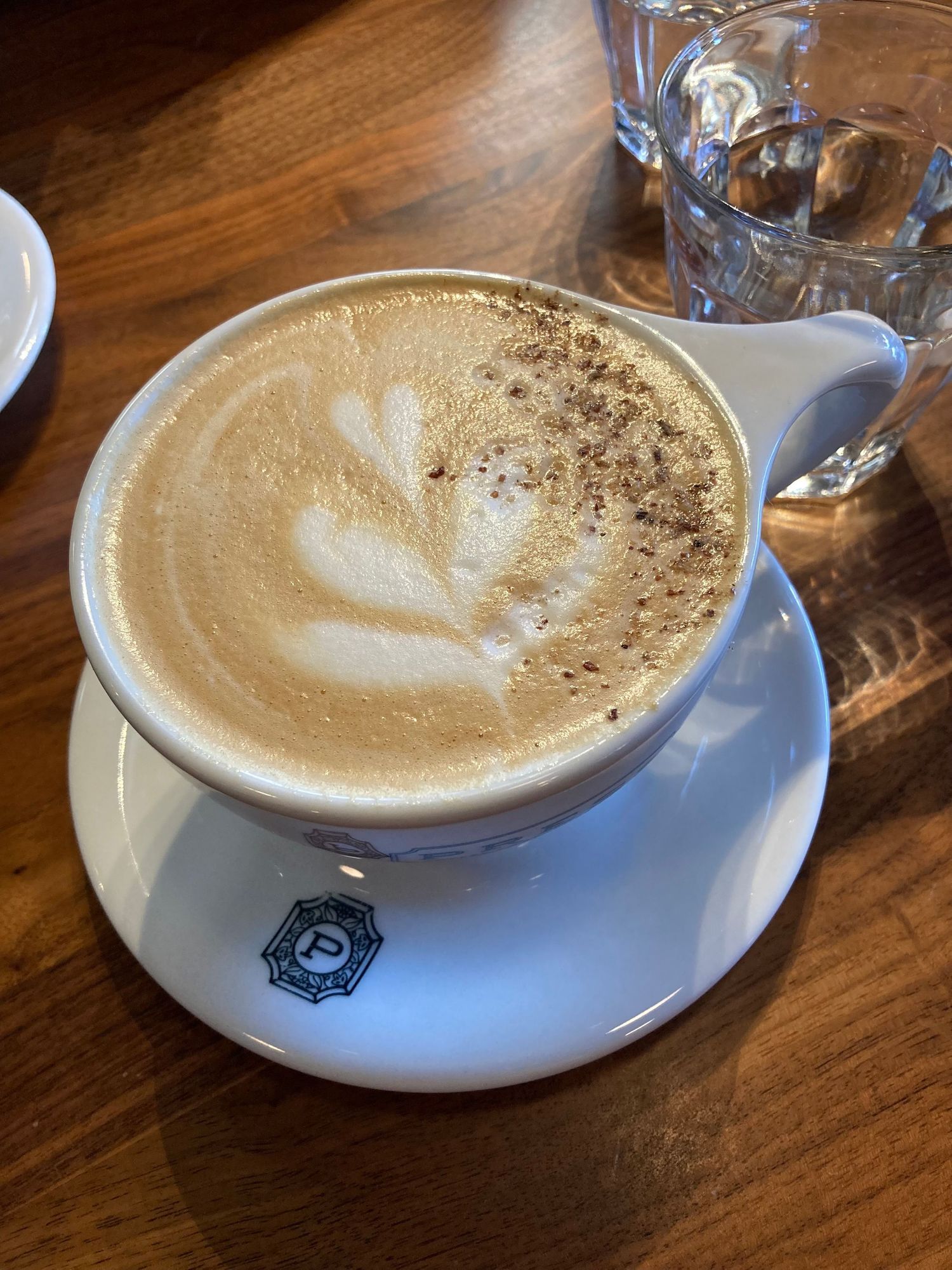 A frothy cappuccino with latte art and cocoa dusting in a white ceramic cup on a saucer on a wooden table.