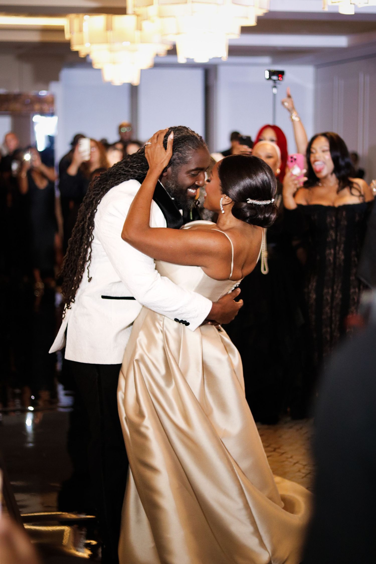 Bride and groom share their first dance at elegant wedding reception, guests celebrating in background.