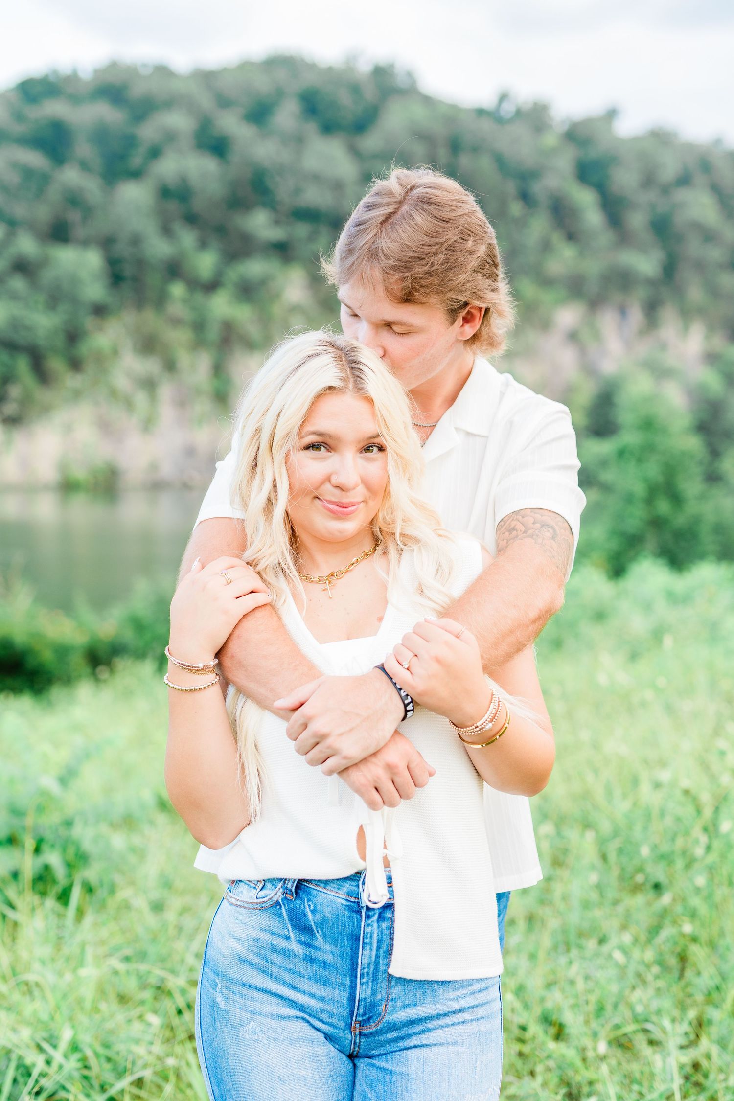 A couple embraces in a sunny meadow with a lake and trees in the background.