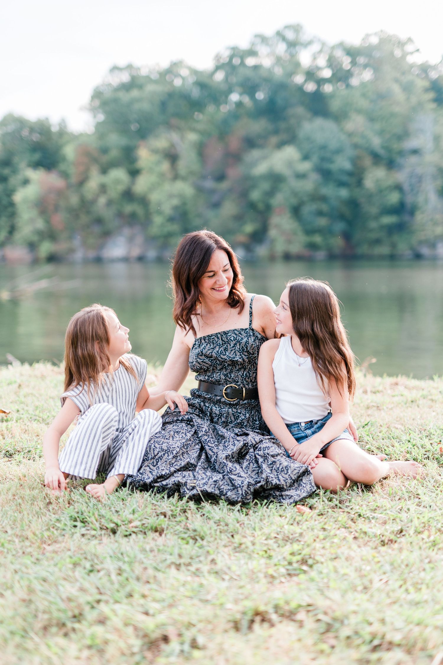 Three people sit together on grass by lake sharing a joyful moment in summer.