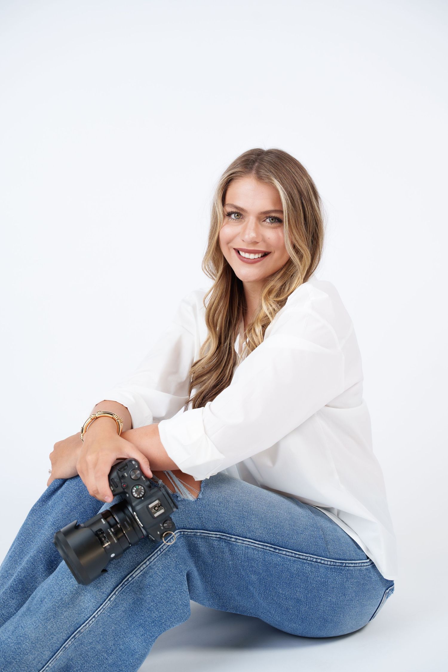 Professional photographer wearing white shirt and jeans sits relaxed with DSLR camera against white background.