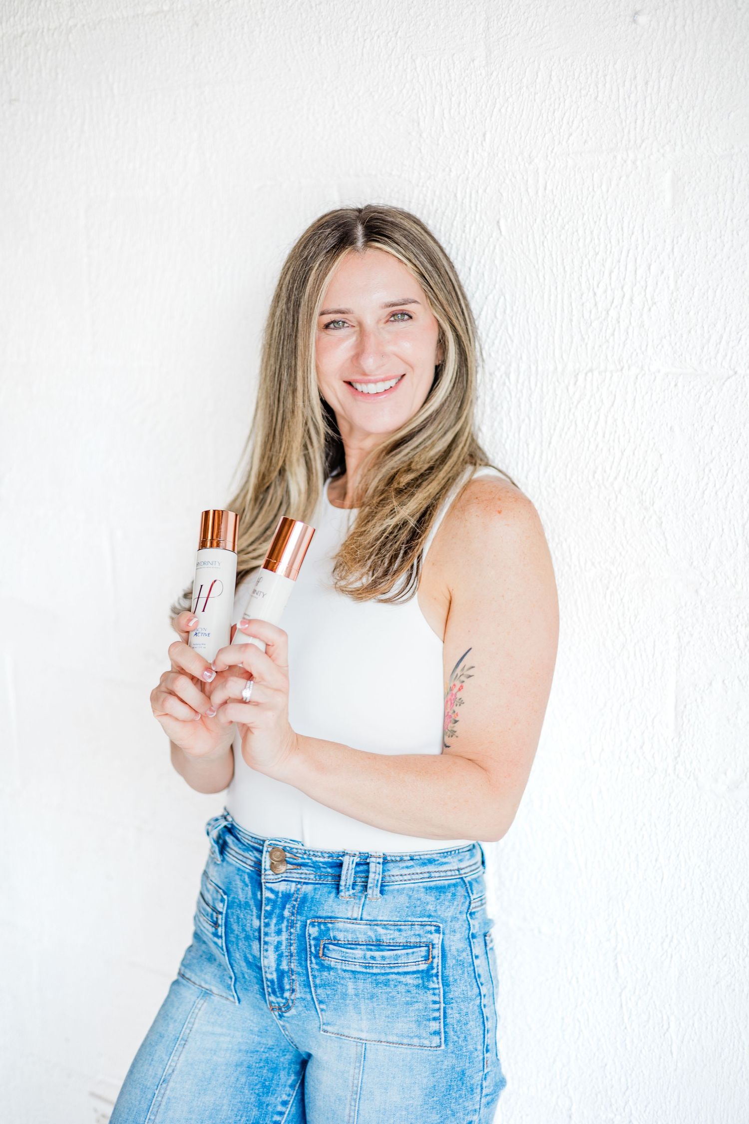 Fashion portrait in white tank top and jeans holding beauty products against white wall.
