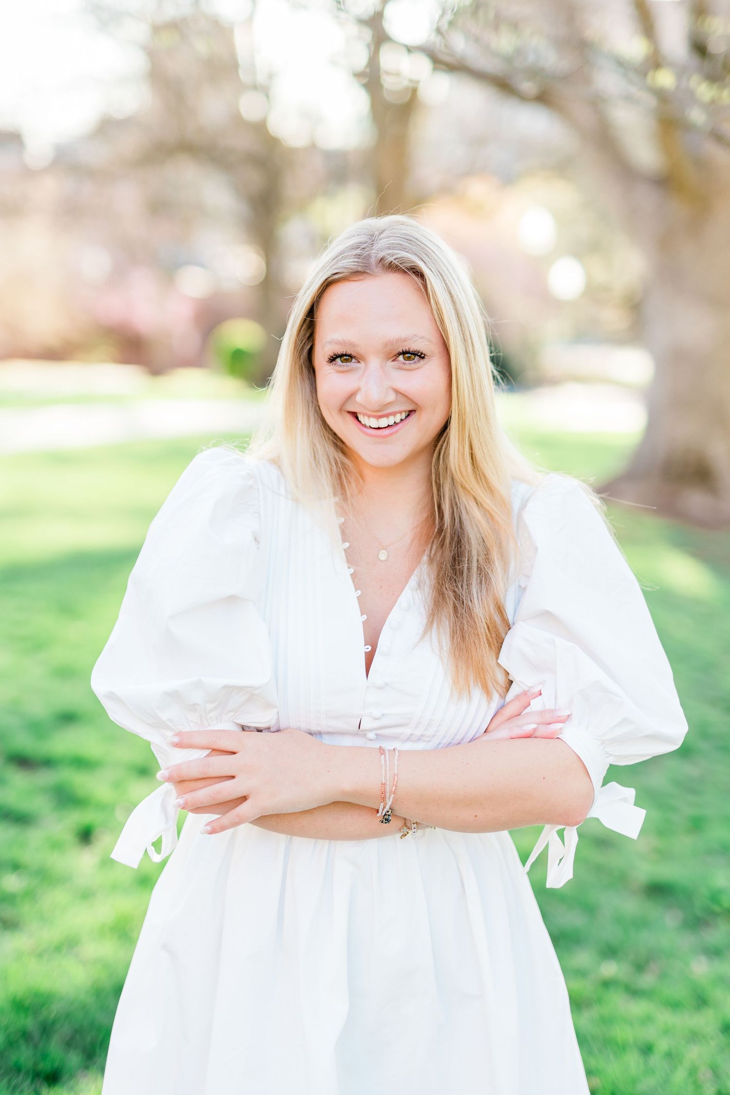 A woman in a white dress with puff sleeves stands outdoors smiling with arms crossed on a sunny spring day.