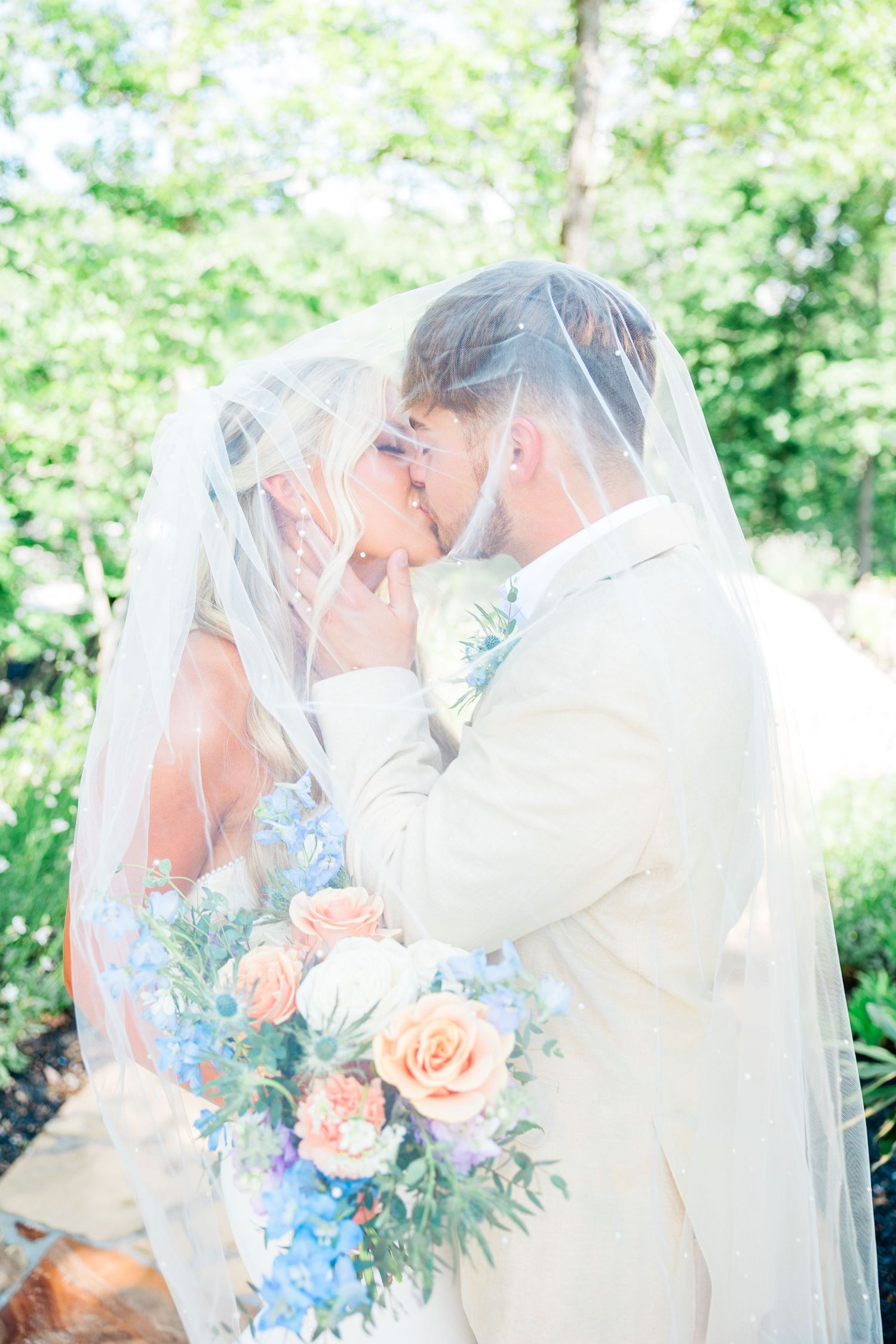 Bride and groom share intimate moments under a flowing veil during outdoor wedding photoshoot with peach roses and blue florals.