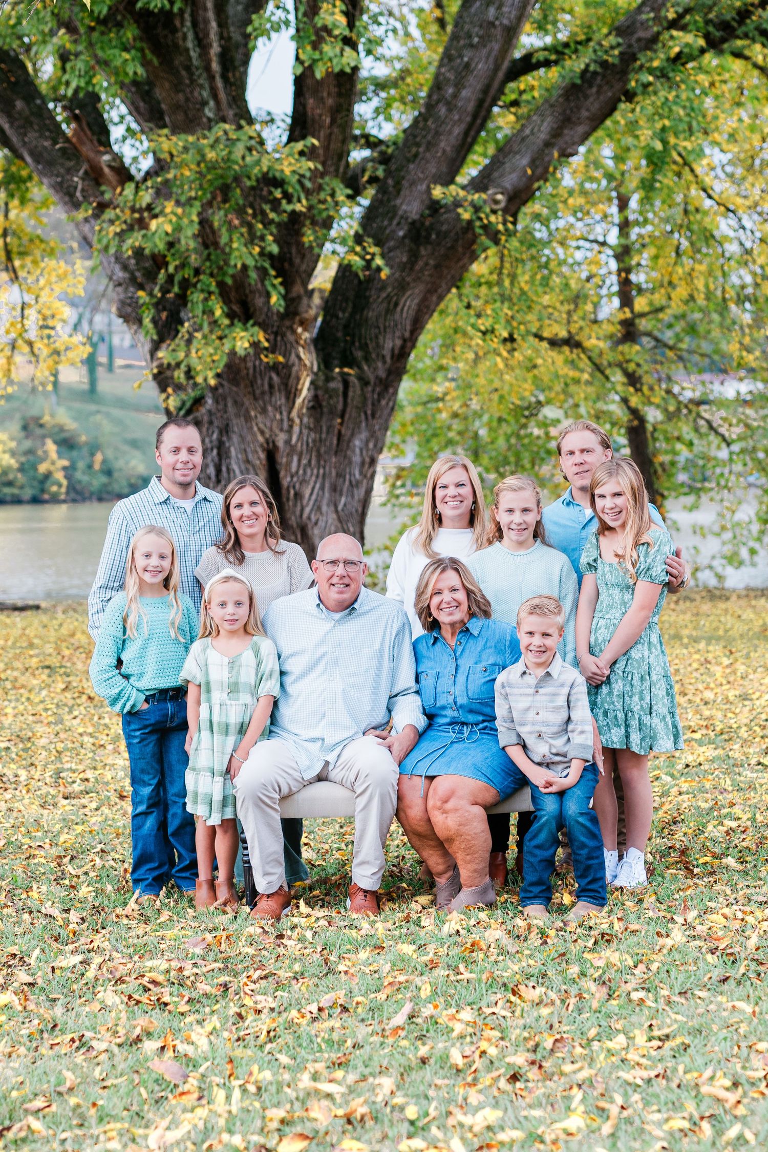 Large family group poses together on autumn grass with majestic tree and lake in background.