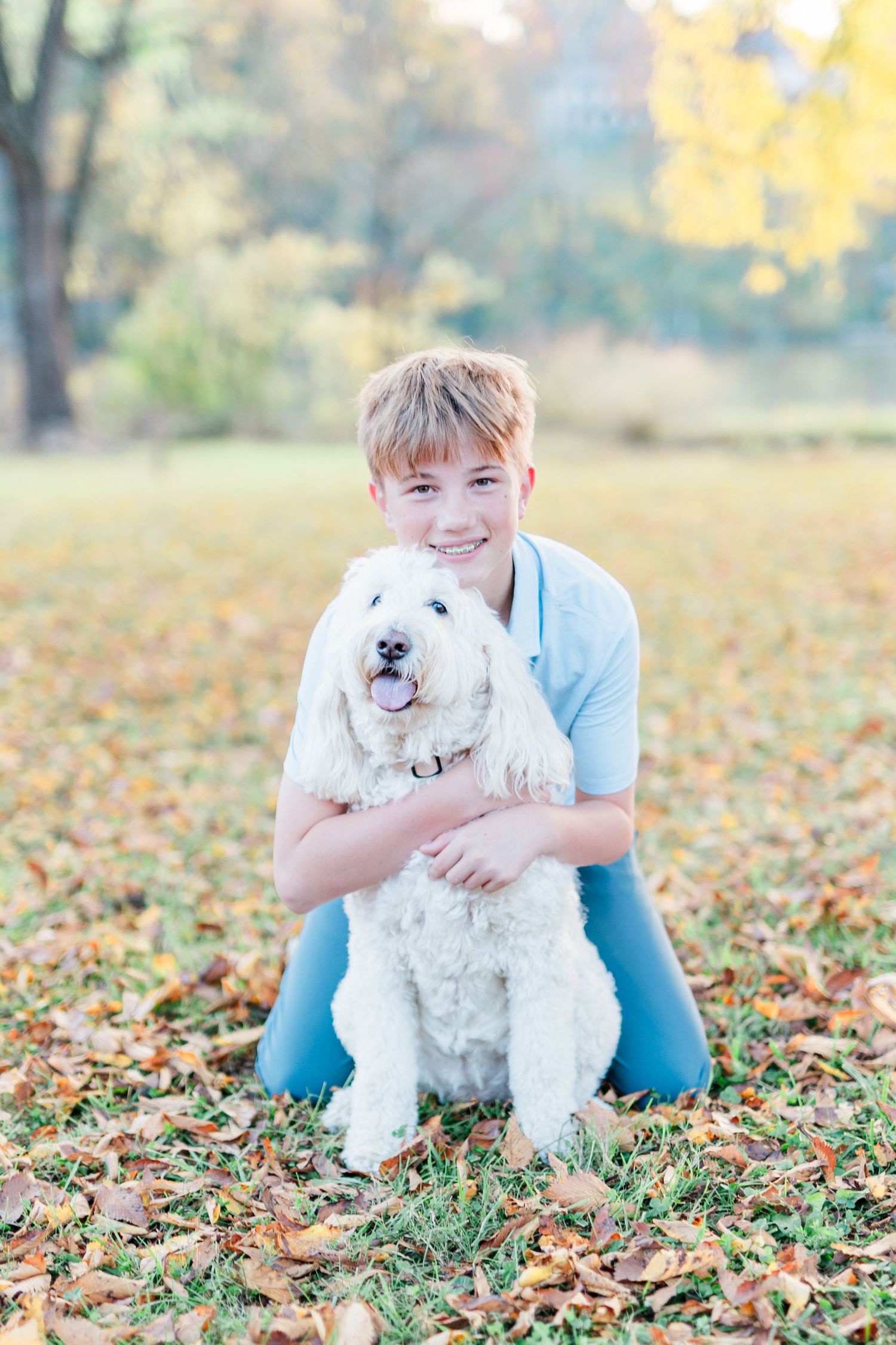 Young person in light blue sweater hugs white fluffy dog in autumn setting with fallen leaves.