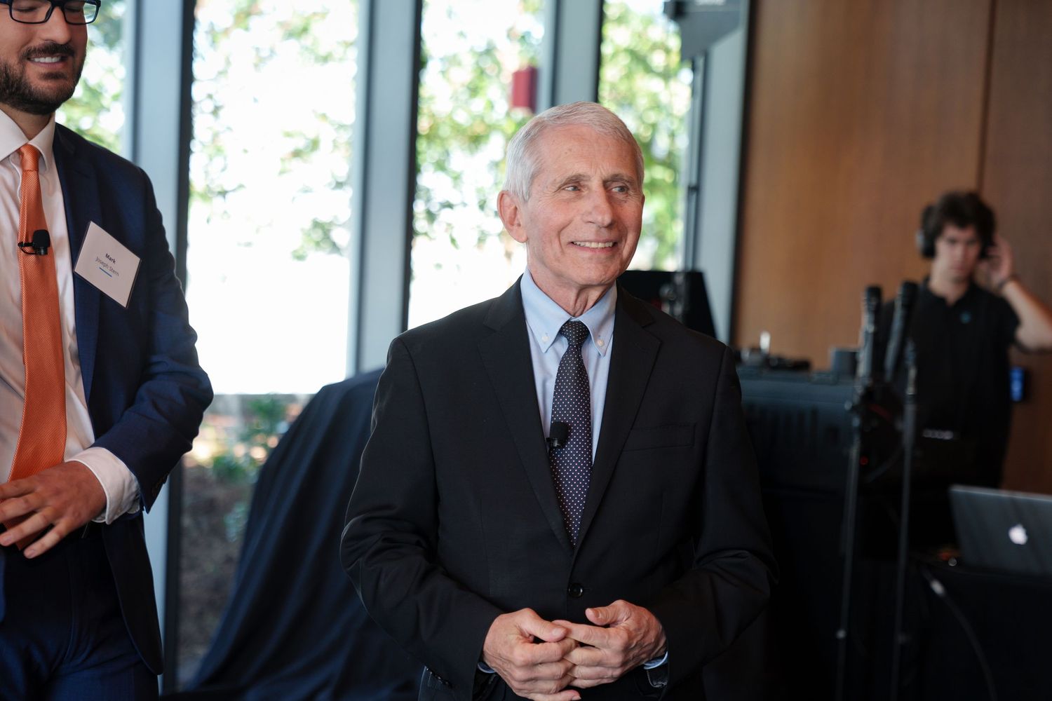 Elderly man in gray suit smiling with clasped hands at a formal indoor event, standing near other attendees.
