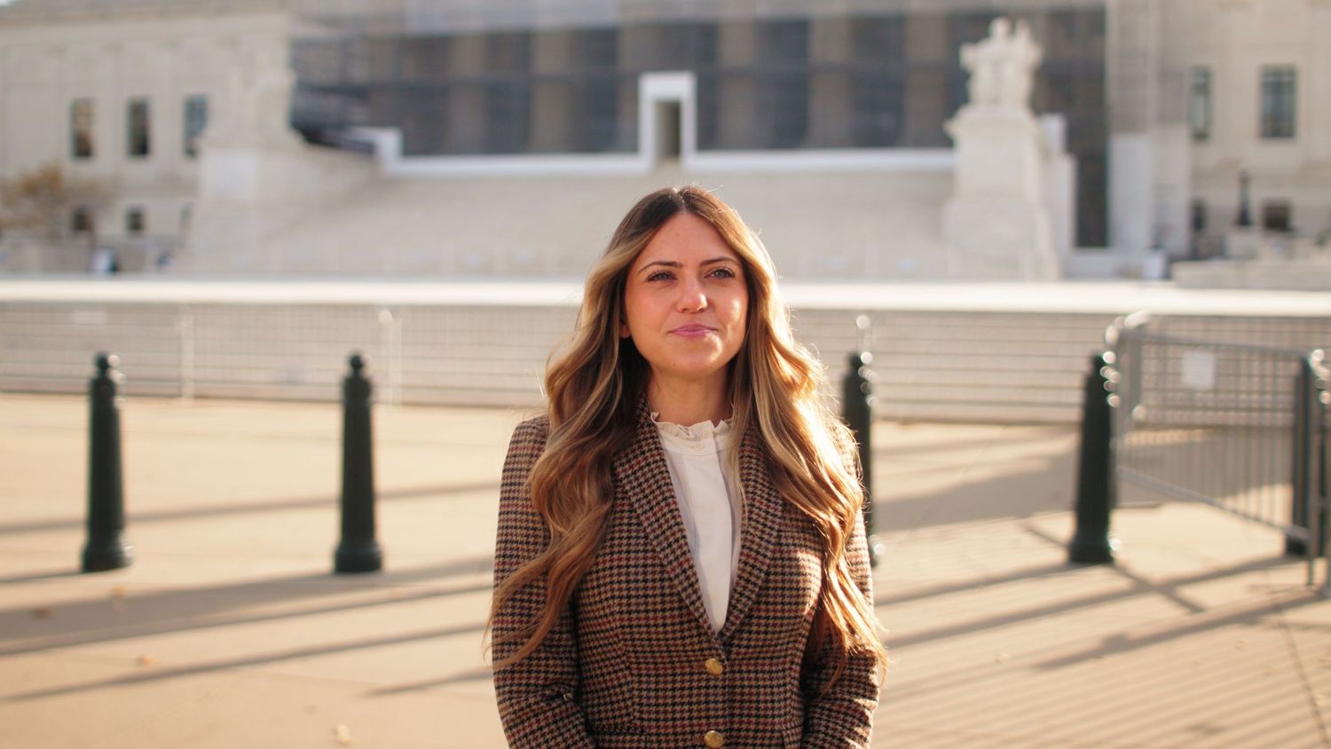 A woman in a brown plaid blazer and white sweater stands outdoors on a sunny day in an urban setting.