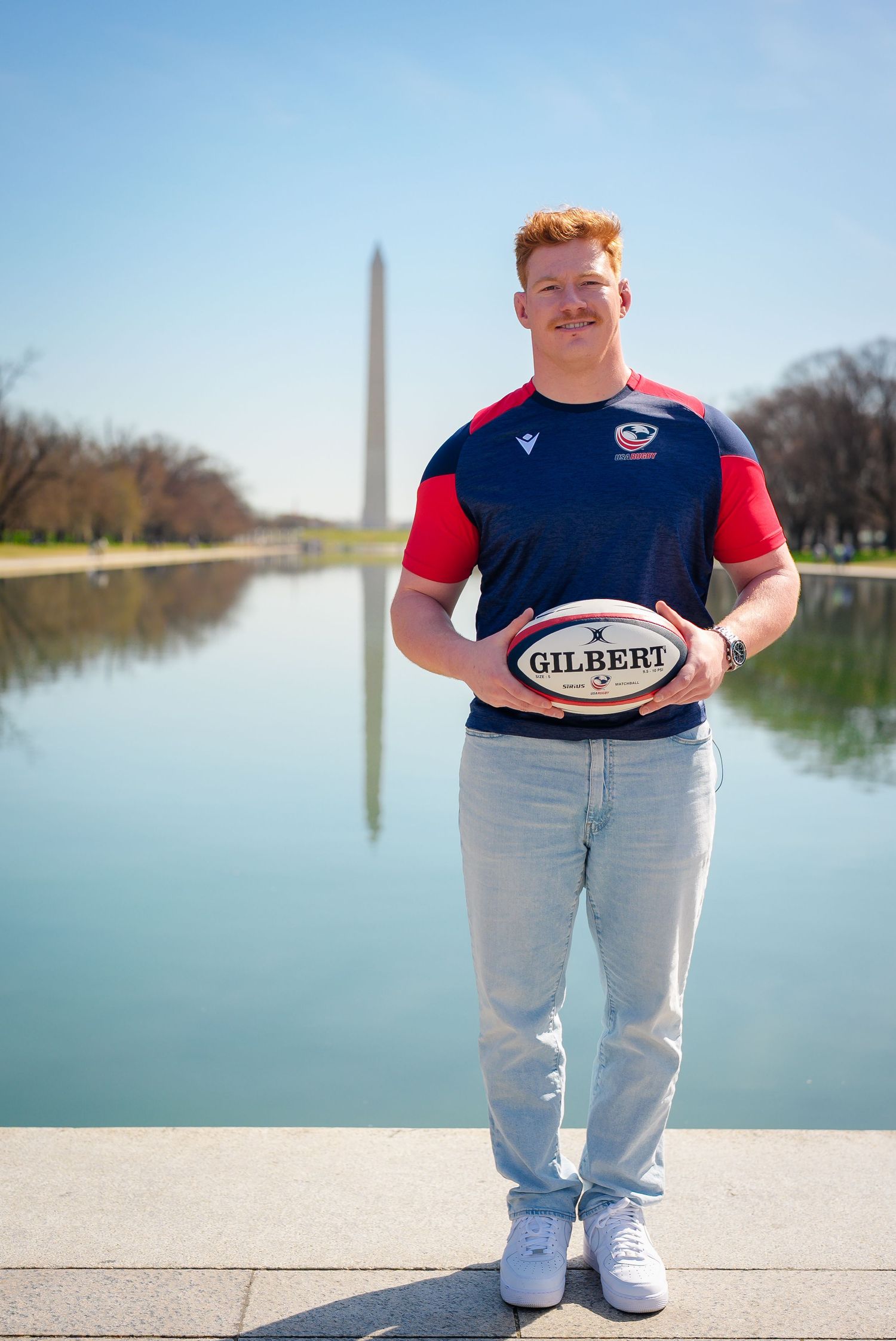 Rugby player poses with Gilbert ball at reflecting pool with Washington Monument in background on sunny day.