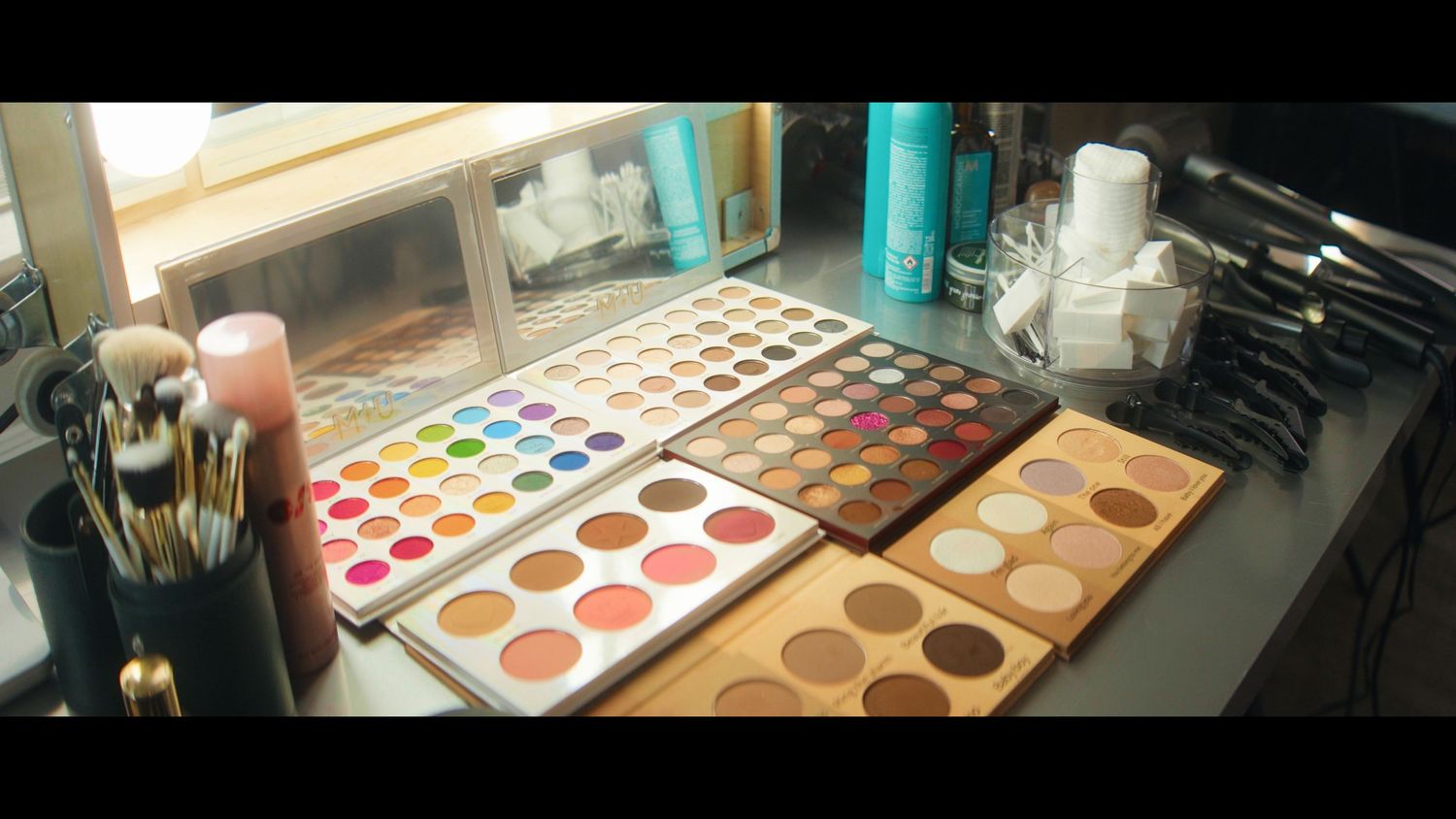 A makeup vanity displays colorful eyeshadow palettes and cosmetic products arranged on a dark surface.